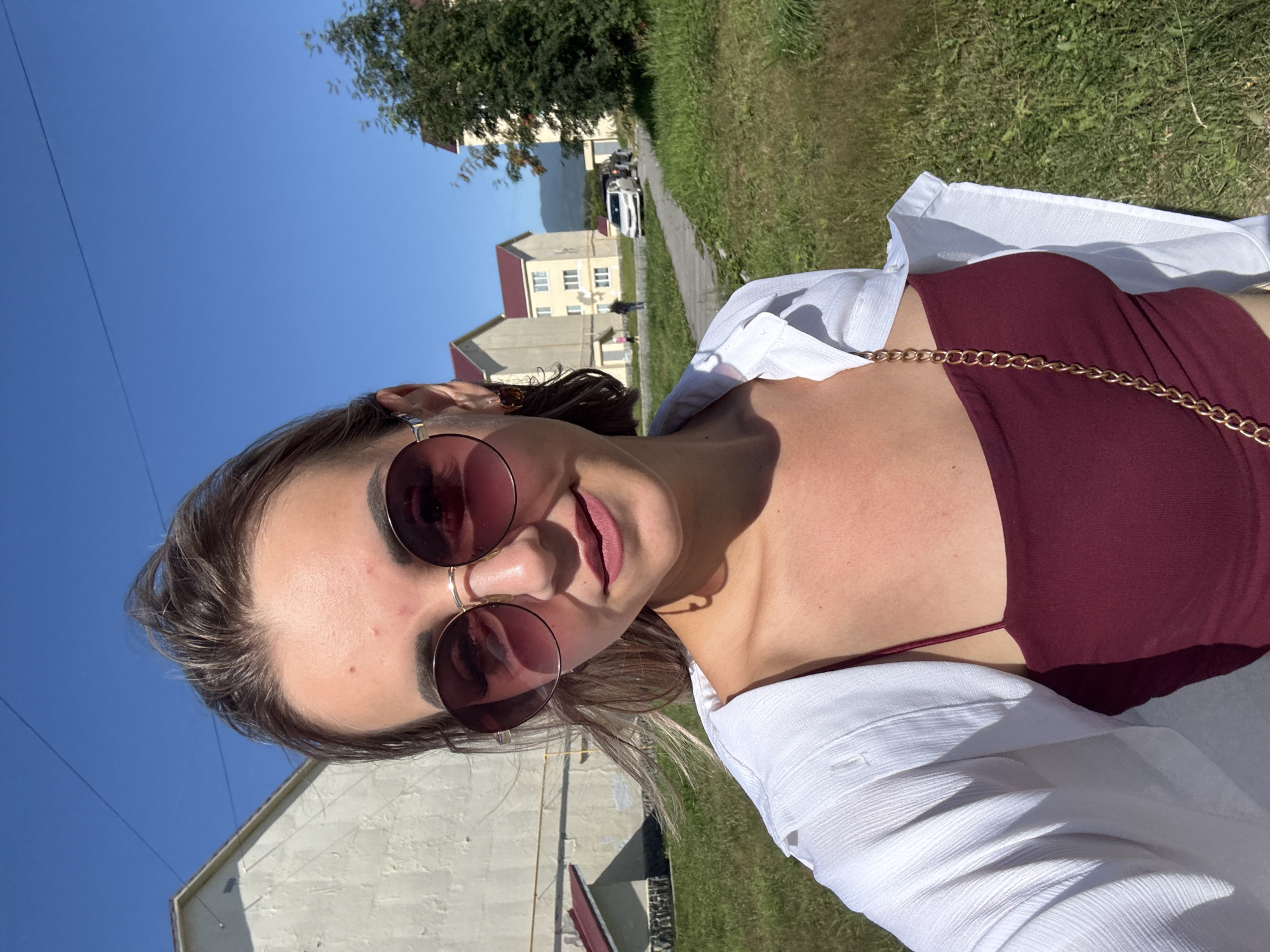 A young Caucasian woman with dark hair and sunglasses takes a selfie outdoors. She is wearing a burgundy top and a white open shirt. A gold chain purse strap is visible. The background features a clear blue sky, green grass, and several buildings. In the distance, a road is visible with two parked cars and a few pedestrians. The overall scene suggests a sunny day in a residential area.