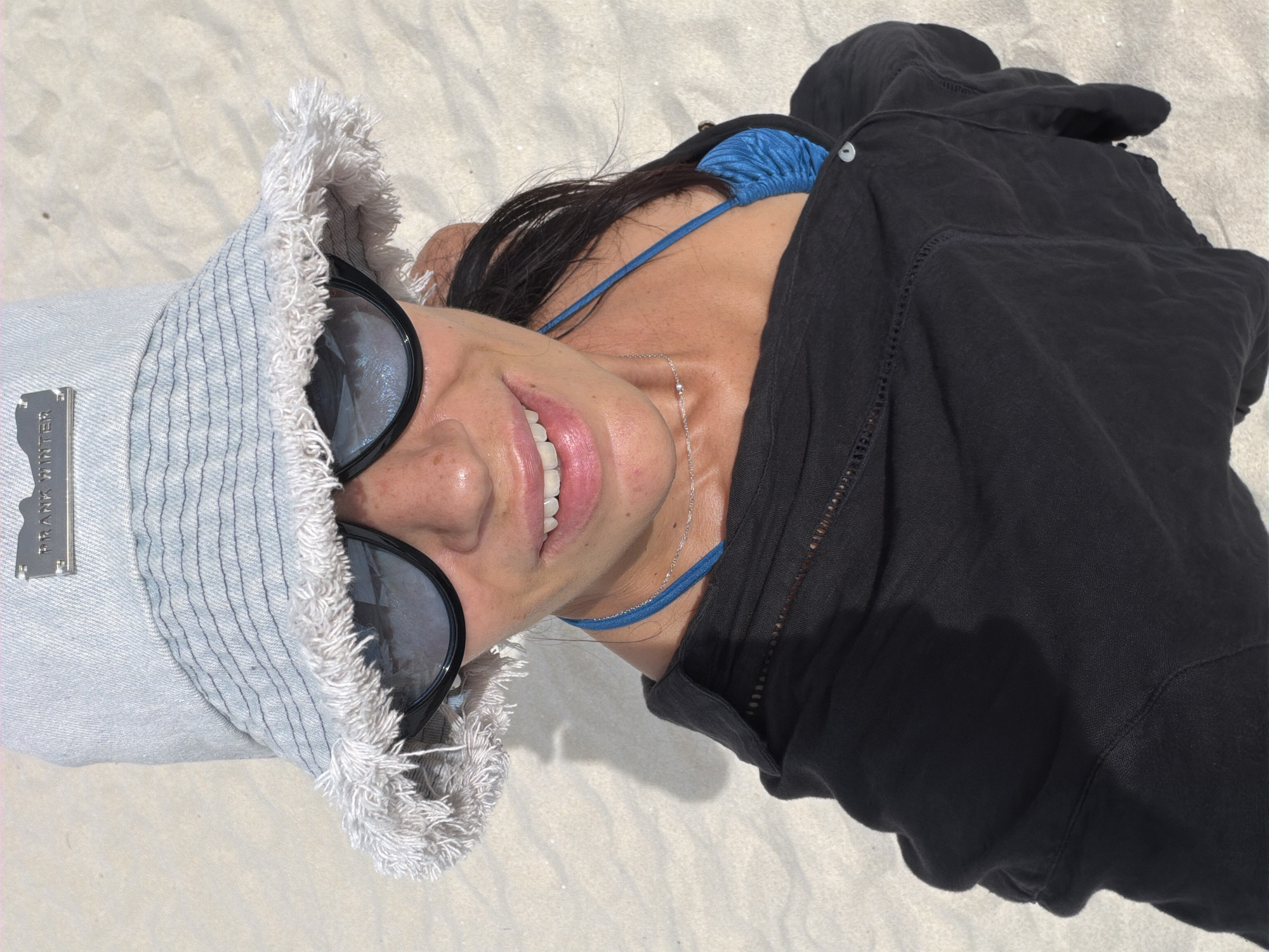 An overhead, medium shot shows a person with dark hair wearing a light blue bucket hat with frayed edges and a silver metal emblem reading "PRANK WINTER." They are also wearing large, dark sunglasses and a black unbuttoned shirt over a blue bikini top. The person is smiling and looking up at the camera, with their face close to the lens. The background is a sandy beach, likely in Baan Chaweng, Thailand, with the texture of fine grains visible. The lighting suggests it is daytime, with a shadow from the person cast on the sand.