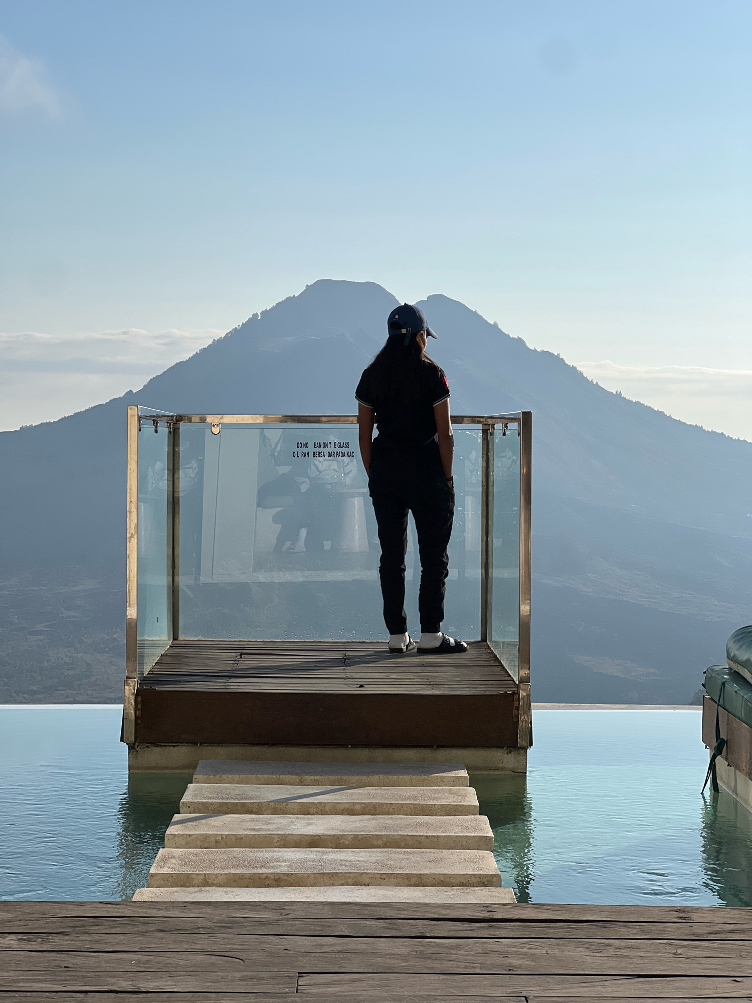 A person stands on a wooden platform overlooking a mountain range in Kintamani, Indonesia. The person is wearing a dark cap, dark shirt, and dark pants, with white socks and dark shoes. They are facing away from the camera, with their hands in their pockets, looking out at the view. In front of the platform is an infinity pool, with stone steps leading into the water. The mountain range is in the background, with a clear blue sky above. The lighting suggests it is daytime, possibly late afternoon or early morning given the long shadows. The scene conveys a sense of tranquility and awe.