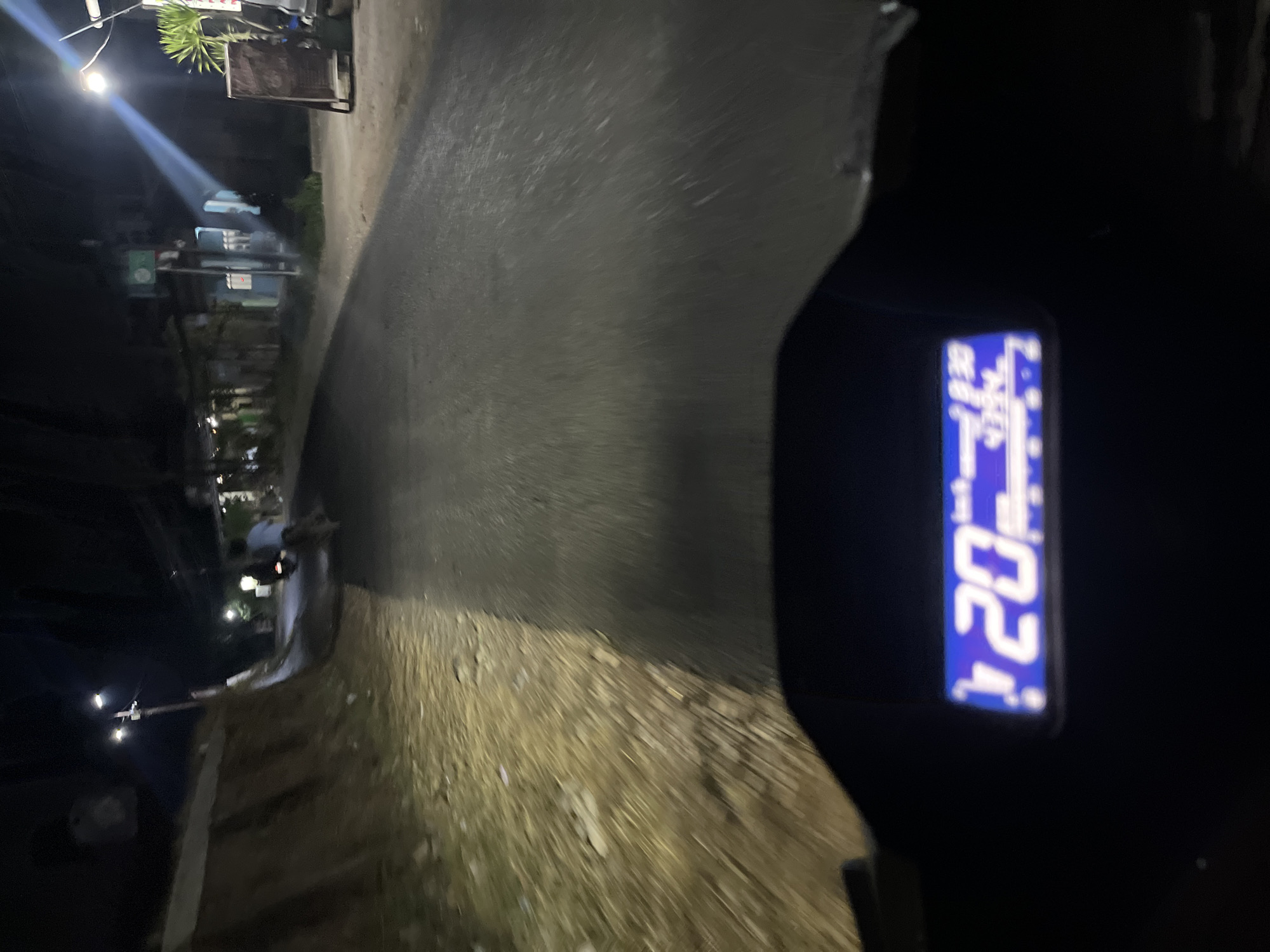 The image is a night-time POV shot from a motorcycle in Karangrandu, Indonesia. The foreground shows a motorcycle's digital display, indicating a speed of 20 km/h and a trip meter of 4394. The road ahead is illuminated by streetlights and the motorcycle's headlights, creating a brightly lit path through the darkness. On the left side of the road, a dirt embankment is visible, illuminated by the motorcycle's headlights. On the right side of the road, there are buildings and businesses with various signs, and a wooden easel-type sign. A street light is visible on a pole, with a beam of light shining down. A person on another motorcycle is visible in the distance, heading away from the camera. The overall atmosphere is that of a typical evening commute on a rural or suburban road.