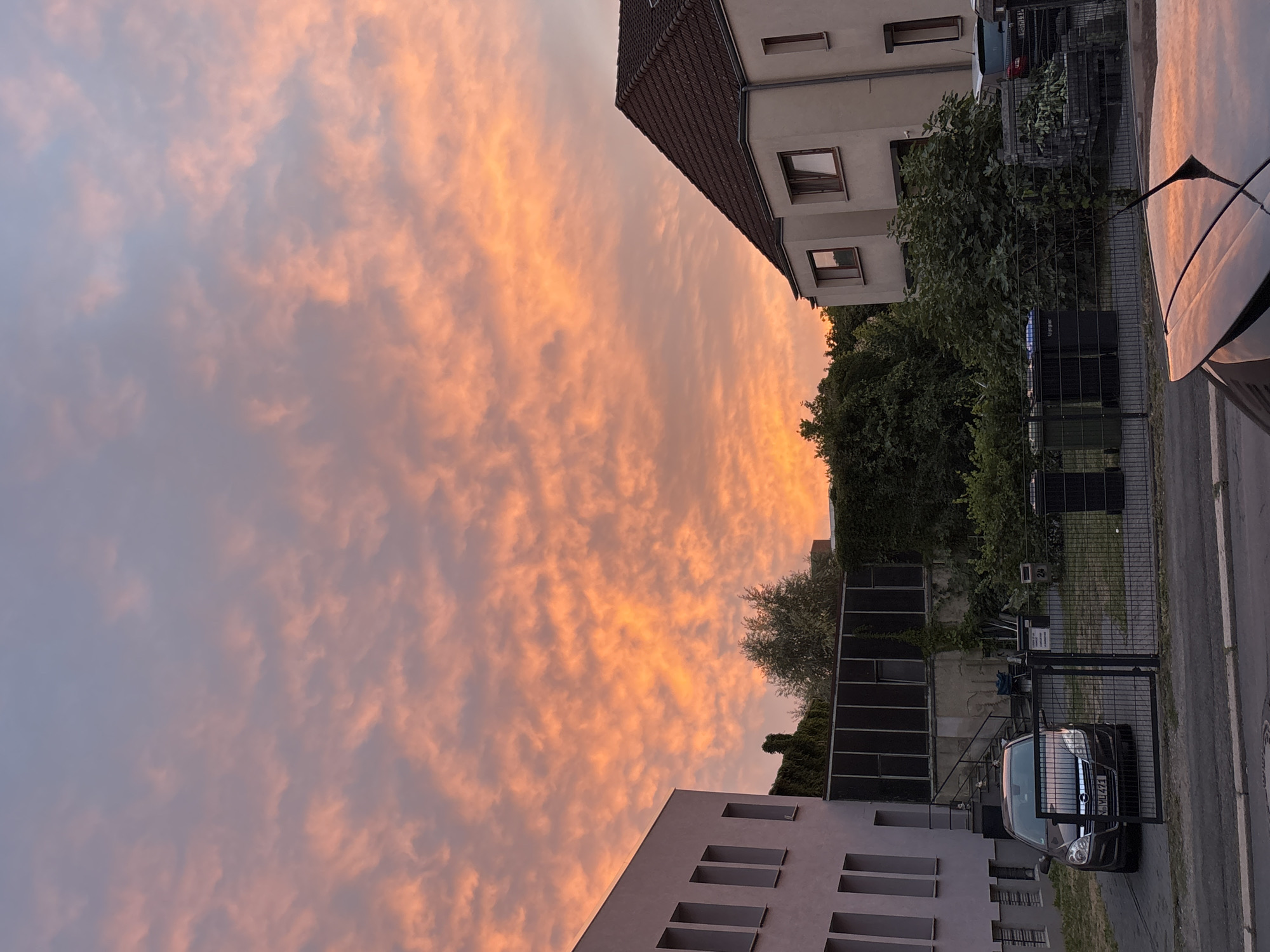 A twilight scene unfolds in Unknown, Unknown, featuring a dramatic sky filled with vibrant orange and pink clouds. Two buildings are visible: a modern, pale-colored structure with rectangular windows on the left, and a light-colored house with a tiled roof and darker-framed windows on the right. Between the buildings, lush green trees and ivy cover a structure. In the foreground, a dark-colored car is parked behind a black metal fence, alongside several large black trash bins. A street runs across the bottom of the image, with the reflection of the colorful sky visible on the car's polished surface.