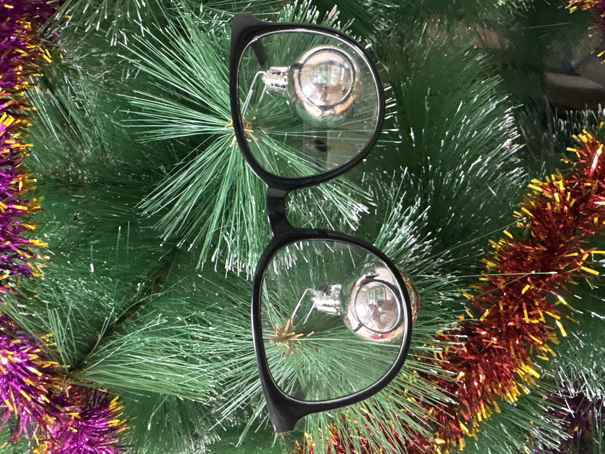 The image shows a pair of black-rimmed eyeglasses lying on top of an artificial Christmas tree. The tree is decorated with silver ball ornaments that are visible through the lenses of the glasses. Tinsel in purple, gold, and red colors is draped around the edges of the tree branches. The location is likely Chaikovsky, Russia, based on the image context being Christmas decorations. No people are visible.