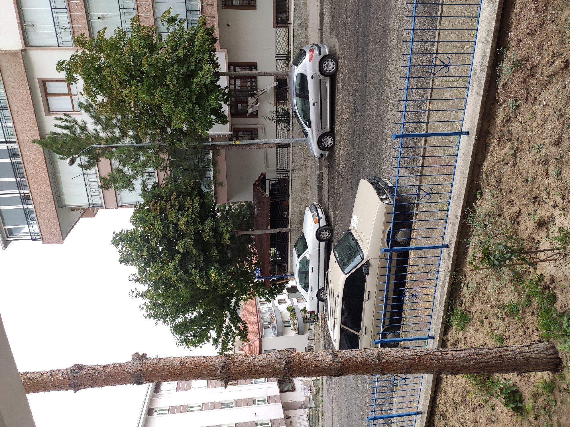 The image depicts a street scene in Mamak, Turkey, with residential buildings and parked vehicles. In the foreground, a light-colored station wagon is parked on the street, partially obscured by a blue metal fence and a tree trunk. Behind it, a white sedan is parked, facing the same direction. Further down the street, a silver hatchback is parked.
The buildings in the background are multi-story residential structures with balconies and windows. There are several trees lining the street, some of which appear to be mature. The sky is overcast. The ground is paved with asphalt and some sections of sidewalk with interlocking pavers. A street sign is visible on a pole, and a lamppost is also present. There are no people clearly visible in the scene, and no discernible actions are taking place. The overall impression is a quiet urban residential area.