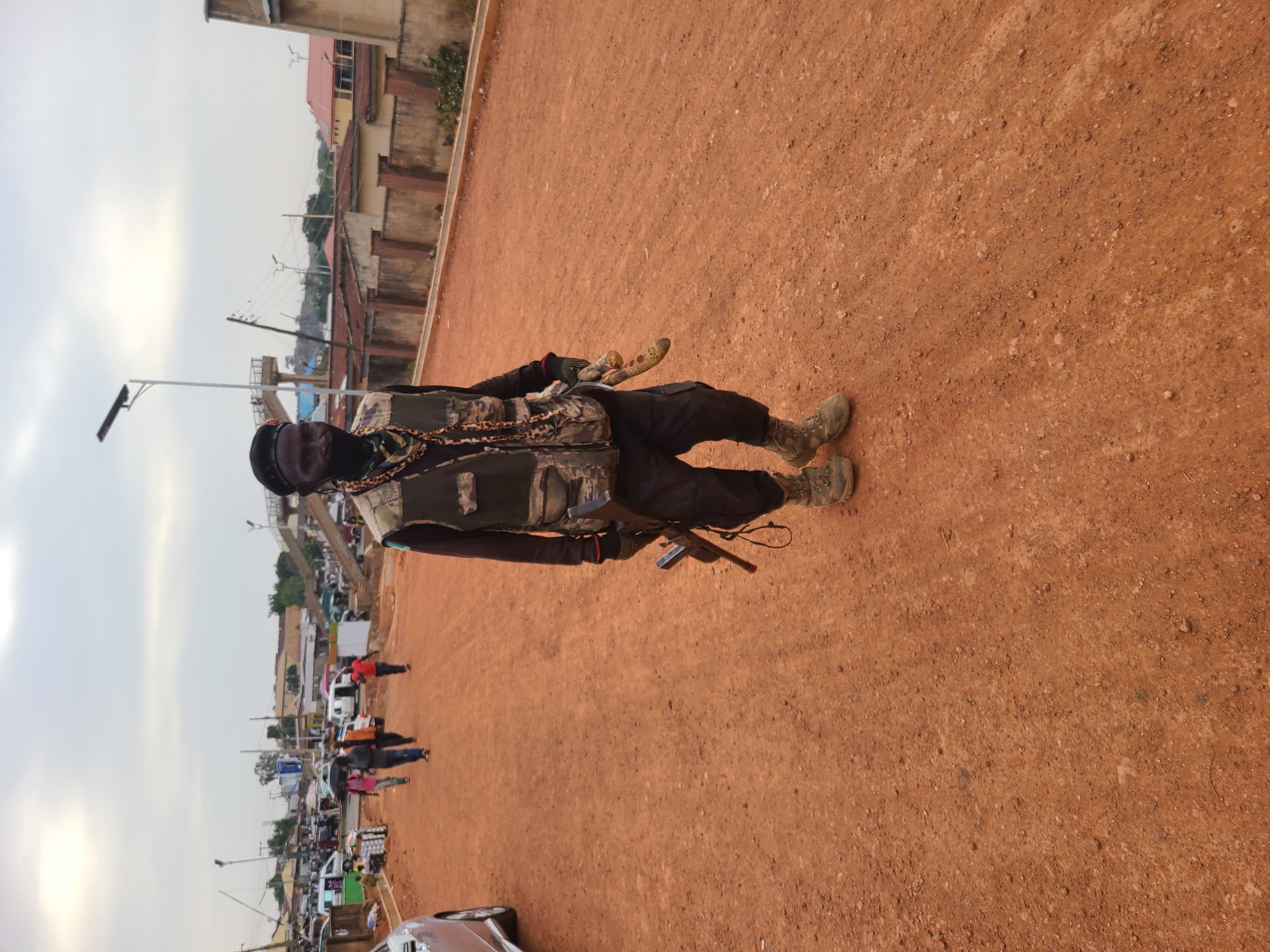 An outdoor, eye-level shot captures a person standing on reddish-brown earth, characteristic of laterite soil, in what appears to be an urban or semi-urban setting in Jos, Nigeria. The main subject, positioned centrally, is a person wearing a dark cap, a dark face covering or balaclava, a camouflage-patterned tactical vest over a dark long-sleeved shirt, dark pants, and military-style boots. They also wear dark gloves and have multiple chains or beaded necklaces draped around their neck and chest.
In their right hand (from the viewer's perspective), the person holds a distinctive, long, curved bladed weapon, resembling a machete or ceremonial sword, with a decorative, possibly yellowish or gold-colored, handle. An object, possibly a holstered pistol or another tool, is visible attached to their left hip.
The background reveals a bustling street scene with several other people walking and various vehicles, including cars and vans, parked or moving. Buildings with different architectural styles, some featuring red roofs, line the street. Utility poles with power lines and streetlights are visible, along with what appears to be an elevated structure or bridge in the distance. The sky is overcast or features diffuse light, suggesting either early morning, late afternoon, or a cloudy day. No legible text is visible in the image.