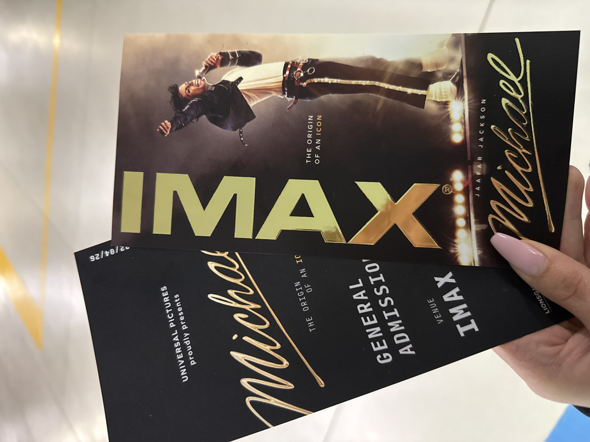 The image shows a person holding a movie poster and a ticket for the "Michael" movie at an IMAX venue. The poster features a picture of Michael Jackson, and the ticket states "General Admission" and "IMAX". The poster also notes that it is "The Origin of an Icon" and that the movie is by Jaafar Jackson. The ticket and poster prominently display the name "Michael" in a cursive gold font. "UNIVERSAL PICTURES proudly presents" is also printed on the ticket. The date 22/04/26 is written on the ticket. The venue is LIONSGATE. The location is likely a cinema due to the movie poster and tickets being held in hand. The location is Sofia, Bulgaria is not specified.