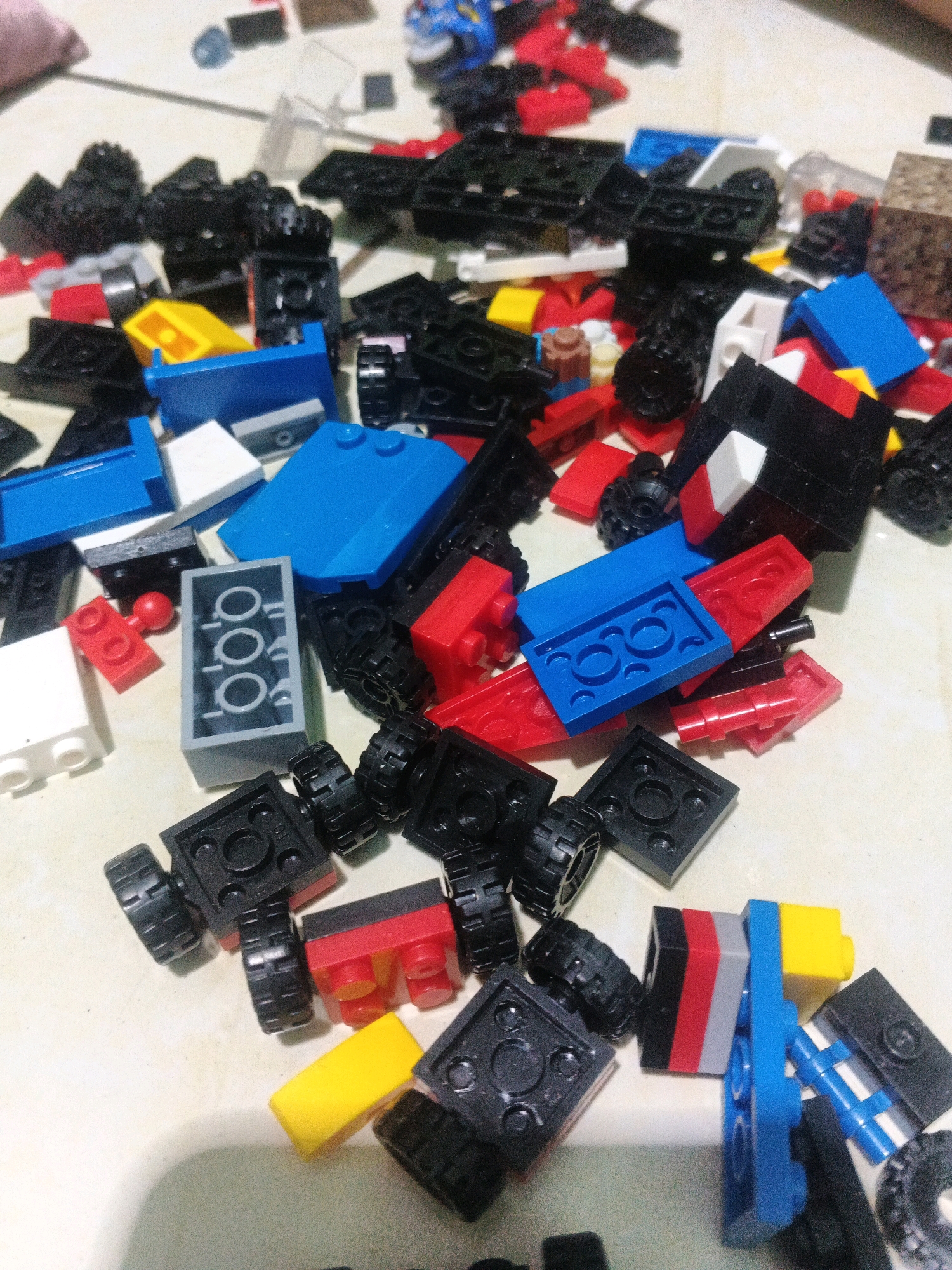 The image displays a collection of colorful LEGO bricks scattered on a light-colored, textured surface. The bricks are predominantly red, blue, yellow, black, and white, with some gray pieces visible. Several of the bricks appear to be parts of vehicles, such as wheels and chassis components. The lighting suggests an indoor environment, likely a tabletop. There is no visible indication of the city of Kadu, Indonesia, or any people.