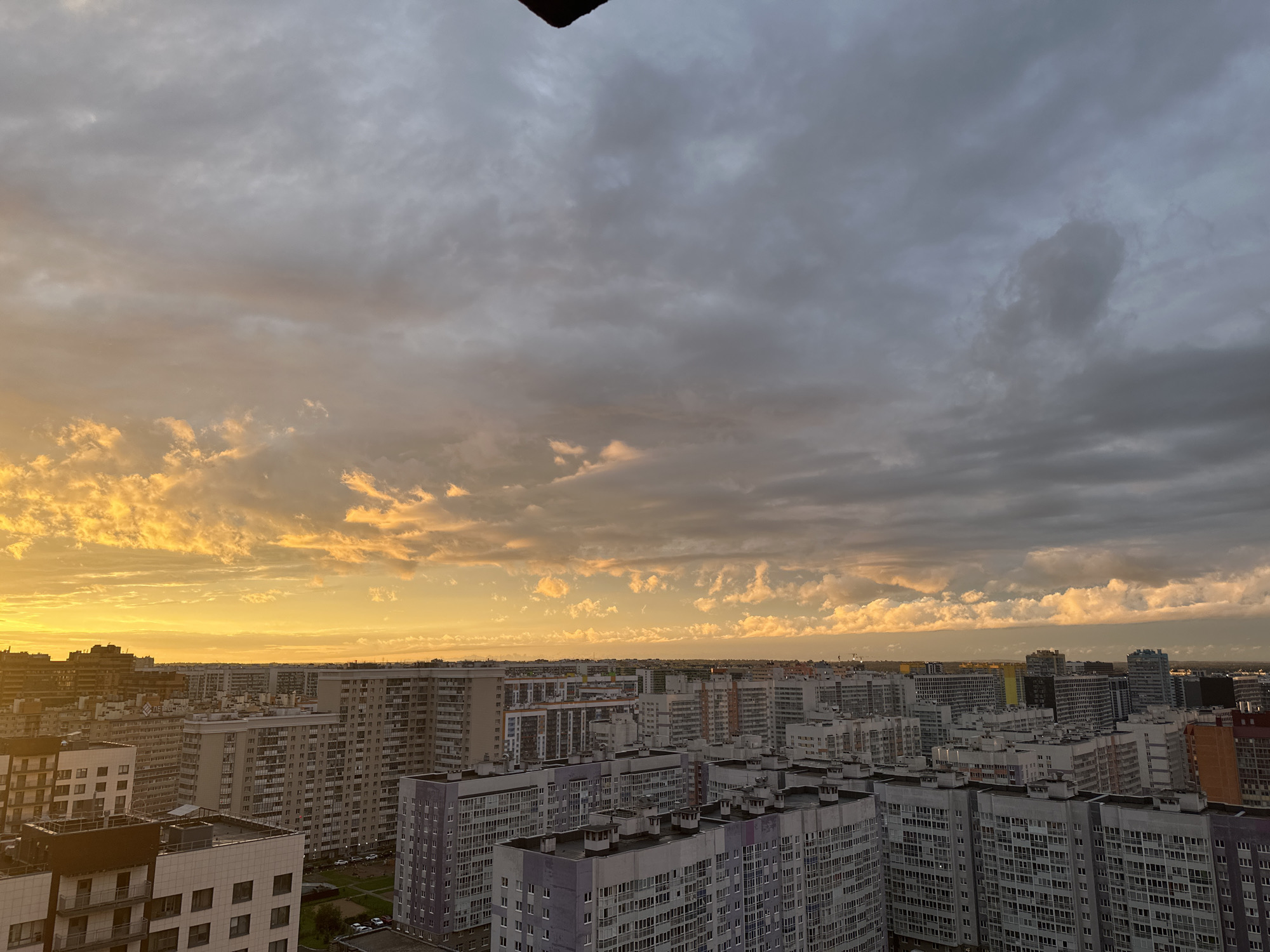 A high-angle, wide shot captures a sprawling urban landscape under a dramatic sunset sky in an undisclosed city in Russia. The foreground is dominated by the rooftops and upper floors of numerous residential apartment buildings, characterized by their modern, stacked designs and uniform window patterns. Some buildings feature balconies, while others have visible ventilation systems or rooftop protrusions. The architecture suggests a densely populated residential area.
The mid-ground continues with more apartment blocks, varying in height and architectural style, forming a dense urban fabric that stretches into the distance. The buildings are predominantly constructed from concrete and painted in muted tones of grey, white, and beige, with occasional splashes of color like orange or yellow on some facades.
The background is defined by the horizon where the cityscape meets the sky. The sky itself is a striking element of the image, featuring a mix of heavy, dark grey clouds interspersed with dramatic streaks of bright orange and yellow from the setting sun. The sunlight illuminates the undersides of some clouds, creating a vibrant contrast against the darker, upper portions of the sky. There is a dark, angled object visible in the upper-left corner, possibly part of a building or structure from which the photo was taken. No people are discernible in the frame, and the scene appears to be static, depicting a typical urban environment at dusk.