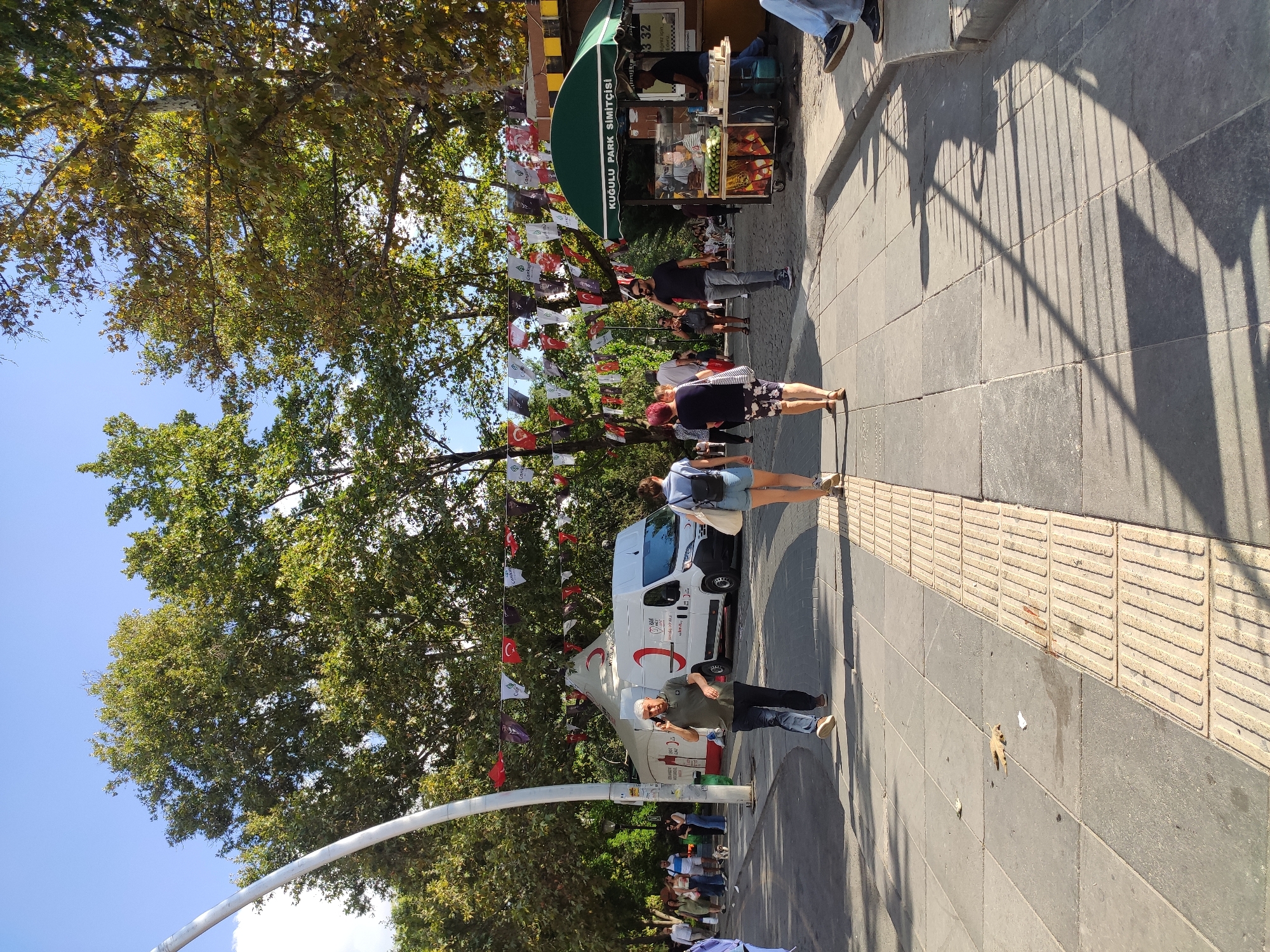 In the city of Ankara, Turkey, at Kuğulu Park, a street scene unfolds on a sunny day. A paved walkway, demarcated by tactile paving for the visually impaired, runs through the park. On the left, a white van with the Red Crescent logo is parked, suggesting a public service or event. A man in a green polo shirt and dark pants walks while talking on his phone, passing by the van. Further down the path, two women walk away from the camera, one in denim shorts and a light blue top, the other in patterned shorts and a dark top. A small dog on a leash is visible near them.

To the right, a green awning shades a food stall with the sign "KUĞULU PARK SİMİTÇİSİ," indicating it sells simit, a Turkish bagel. Several people are gathered around the stall, including vendors and customers. The background is filled with lush green trees, their leaves catching the sunlight, and a clear blue sky overhead. Small triangular flags, some bearing Turkish flag colors and symbols, are strung across the area, adding a festive atmosphere. The overall scene depicts a casual public space in Ankara, likely a popular spot for recreation and local commerce.