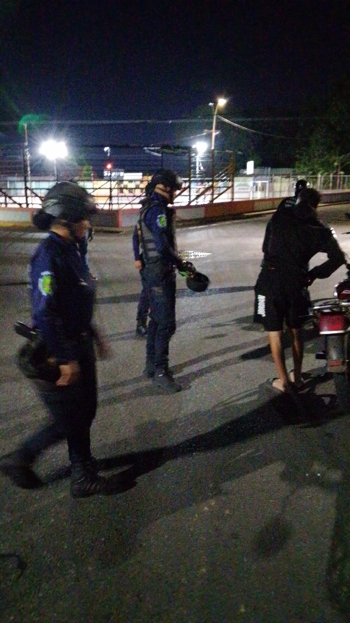 The image depicts an outdoor scene at night in Tinaquillo, Venezuela. Three individuals, likely law enforcement officers based on their uniforms and tactical gear, are visible. One officer is walking toward the left side of the frame, while the other two stand near a person dressed in black clothing and what appears to be a parked motorcycle. The environment includes an asphalt surface, street lighting, and a fence or barrier in the background.