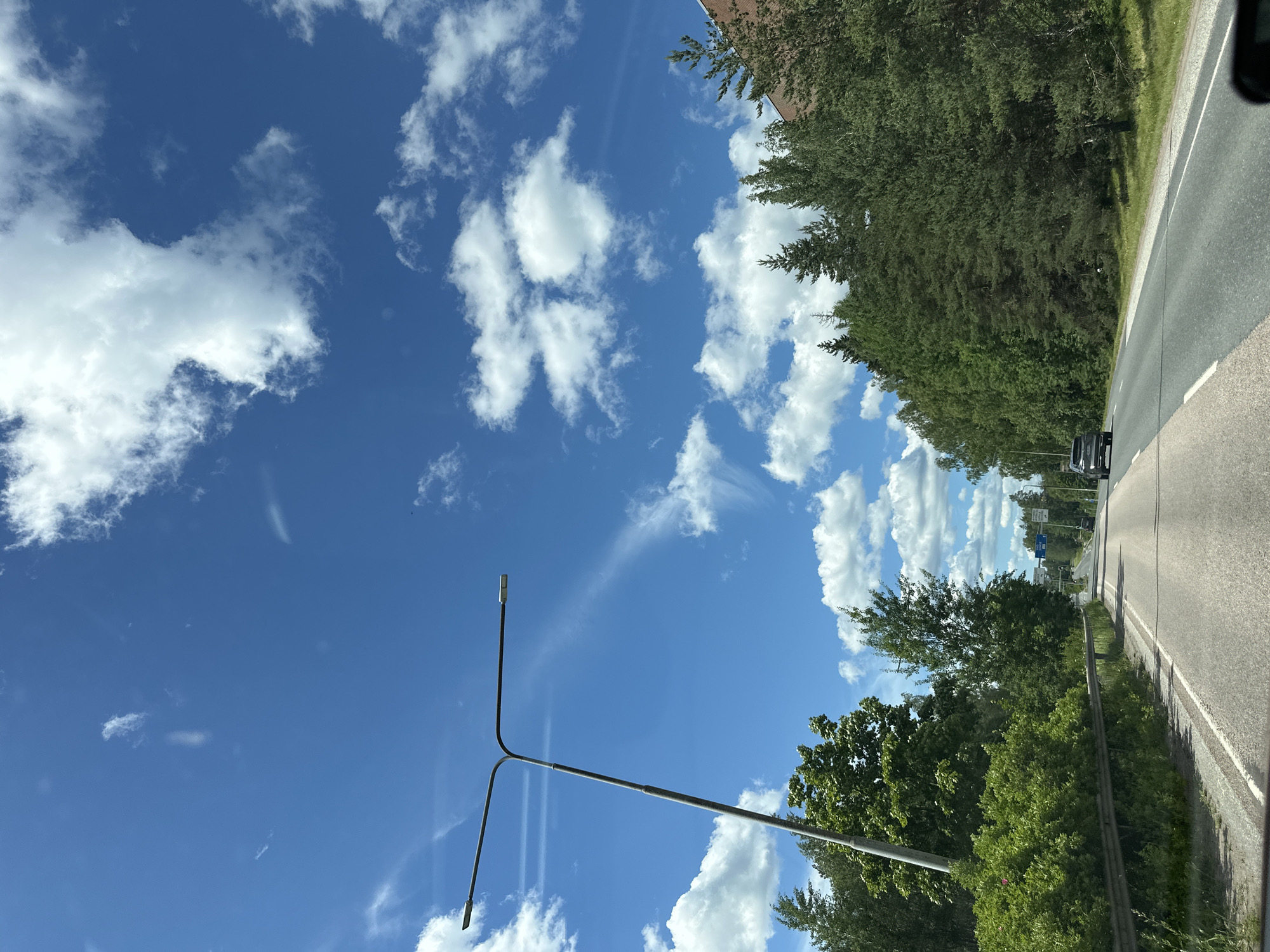 This is a vertical, eye-level shot of a bright blue sky with fluffy white clouds. The sky takes up most of the frame, with the sun not directly visible but implied by the bright illumination and shadows. The weather appears to be fair and sunny.
On the right side of the image, a paved road runs diagonally from the bottom right corner towards the top of the frame, disappearing into the distance. Trees line the right side of the road, with lush green foliage indicating a warm season. A dark-colored car is visible on the road in the distance. There is also a low metal barrier running along the edge of the road next to the trees.
In the lower left quadrant of the image, a modern street lamp with a unique Y-shaped design is prominently featured. It has two lights at the end of its arms, pointing in different directions. The street lamp is silhouetted against the bright sky.
The overall scene suggests a drive or a journey on a sunny day through a somewhat rural or suburban area with ample greenery. There is no visible text in the image.