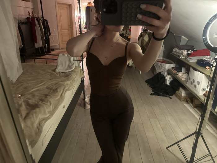 A woman in a brown, form-fitting outfit stands in front of a mirror, taking a selfie with her phone obscuring her face. She wears a brown bodysuit with thin straps and matching high-waisted pants. The room features a bed with a tan blanket and a clothing rack with various garments. In the background is a shelf with items stacked on it, and a ring light on a stand is visible. The floor is light-colored wood.