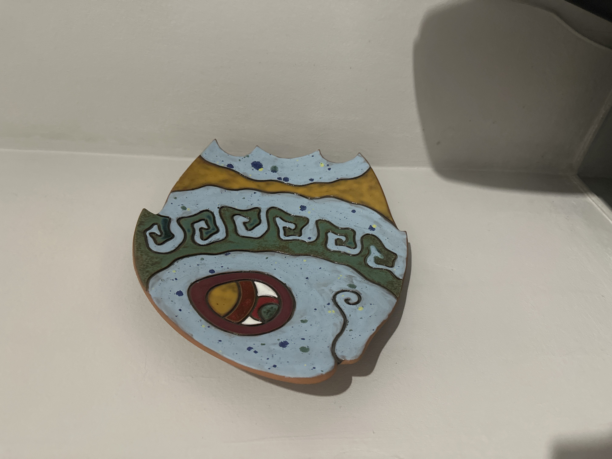 A ceramic wall hanging in the shape of a stylized fish is mounted on a light-colored wall. The fish has a tan border and is decorated with blue, green, and brown patterns. There is a circular "eye" design with concentric rings of brown, red, and white on the lower part of the fish. The top edge of the fish has a wavy, serrated design. The location in Yerevan, Armenia, is not discernible from the image.
