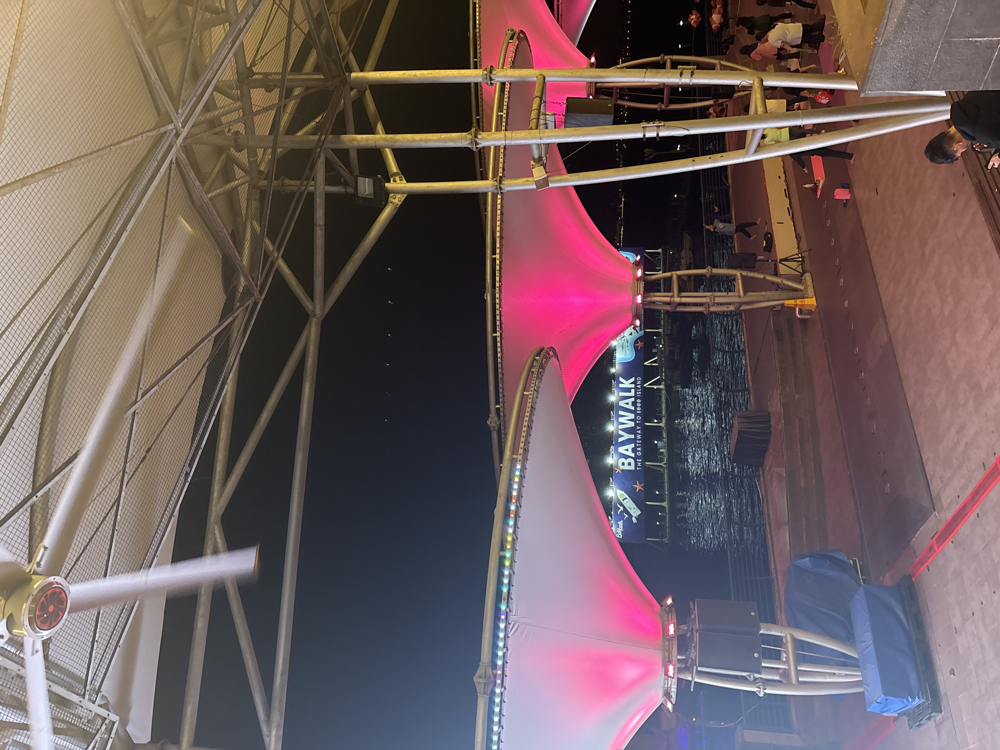 This is a nighttime outdoor scene, likely in North Jakarta, Indonesia, given the location context. The image captures a large, illuminated structure, possibly part of a ferris wheel or a similar amusement ride, with large, white, sail-like canopies that are lit with vibrant pink and colorful LED lights. These canopies are supported by a complex metal framework.
In the background, a body of water is visible, reflecting the lights from the structure and the surroundings. A blue banner with white text that reads "BAYWALK THE GATEWAY TO 1000 ISLAND" is prominently displayed. The text "Baywalk" is also visible in a smaller logo. This suggests the location is the Baywalk Mall or a similar waterfront area known for its views and attractions.
Several people are present in the scene. Some are seen walking or standing on a walkway or platform near the water, while others are gathered on a larger tiled area, some of whom appear to be participating in a yoga or exercise session, with yoga mats visible on the ground. The overall atmosphere appears to be lively and engaging, with the colorful lights and the presence of people suggesting an event or a popular public space. The time of day is clearly night, indicated by the dark sky and artificial lighting. The weather appears to be clear.