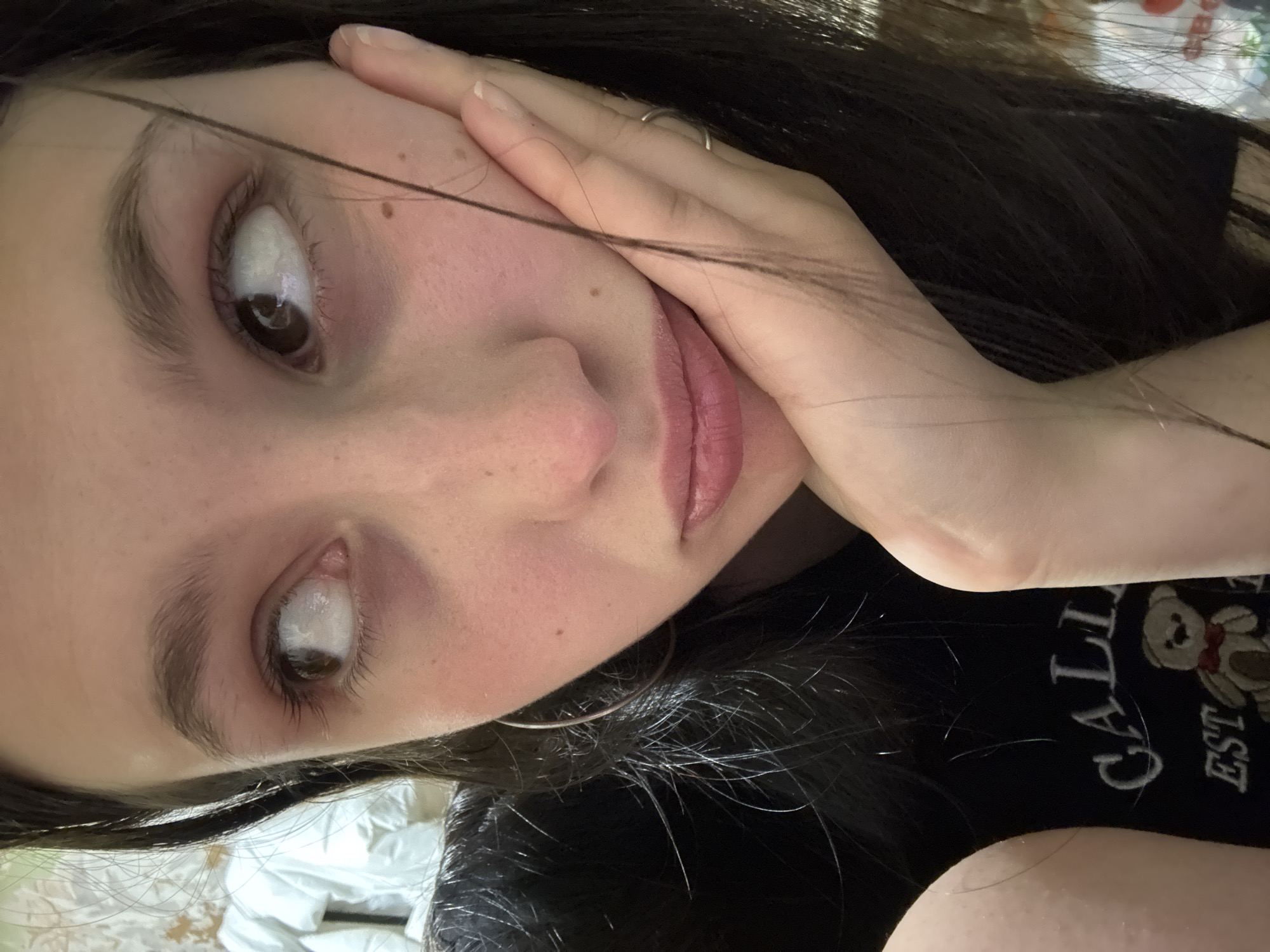 A young woman with dark hair and brown eyes is positioned closely in the frame, her face tilted. Her right hand is raised, her fingers resting gently against her cheek. She is wearing a black t-shirt. The background is blurred. No information indicates the location is in Фоки, Russia.