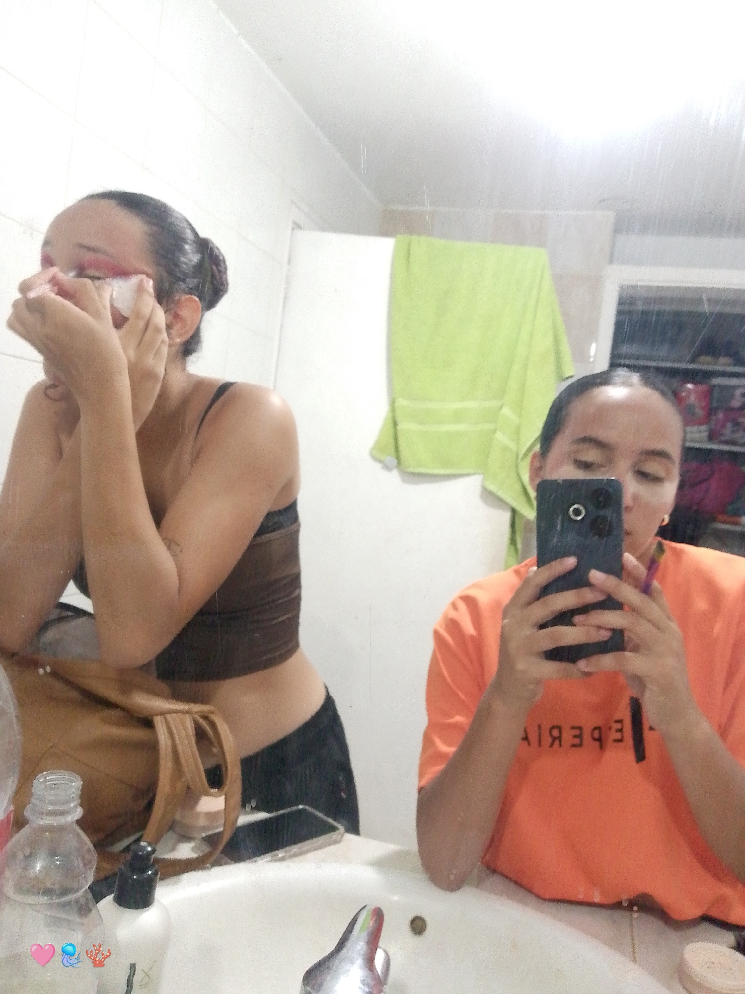 This is a photograph taken in a bathroom, likely in a residential setting in Playa El Agua, Venezuela. The image captures two young women, each appearing in a mirror reflection.
In the left half of the frame, a woman is seen applying makeup around her eyes. She has her hair pulled back into a bun and is wearing a dark brown crop top and black bottoms. To her left, a brown handbag is placed on the counter, and in front of it are various bottles of toiletries.
In the right half of the frame, another woman is holding up a smartphone to take a selfie. She is wearing an orange t-shirt with lettering visible on the back, which appears to read "EXPERIENCIA" in reverse. Her hair is also pulled back. Behind her, a light green towel is hanging on the wall, and shelves filled with items are visible in the background. The overall environment suggests a private space, possibly a home bathroom.