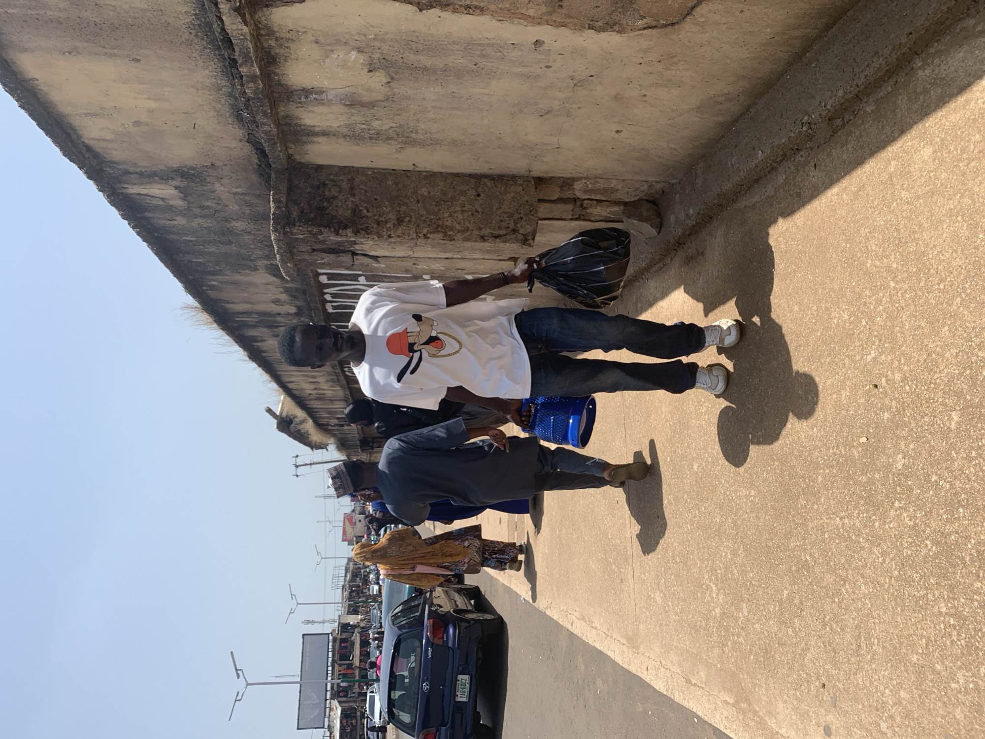 In a street scene in Jos, Nigeria, a man in a white t-shirt and jeans holds a black plastic bag. He stands on a concrete sidewalk beneath an elevated concrete structure, casting a shadow on the ground. Another person, wearing a dark blue tunic and a patterned head covering, walks past with a blue bucket. In the background, a busy street is visible with parked cars and people. The weather appears to be clear and sunny, suggesting it is daytime. The scene captures a moment of daily life in a bustling urban environment. There is text on the license plate of the blue car in the background, "TAQ 60CZ".