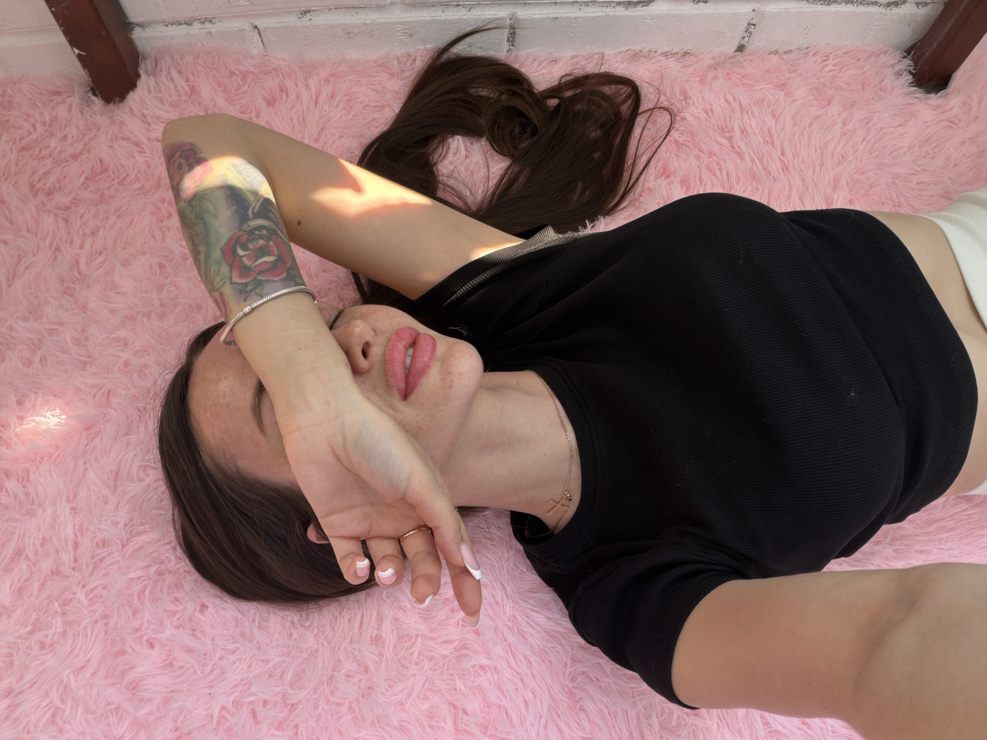 A young woman is lying on a pink, furry rug. She is wearing a black shirt and white shorts. Her left arm is raised, covering part of her face. She has a tattoo on her left forearm and a ring on her left hand. Her hair is dark brown and spread out on the rug. A gold necklace is visible around her neck. The background shows a white brick wall. The location is not Saint Petersburg, Russia.