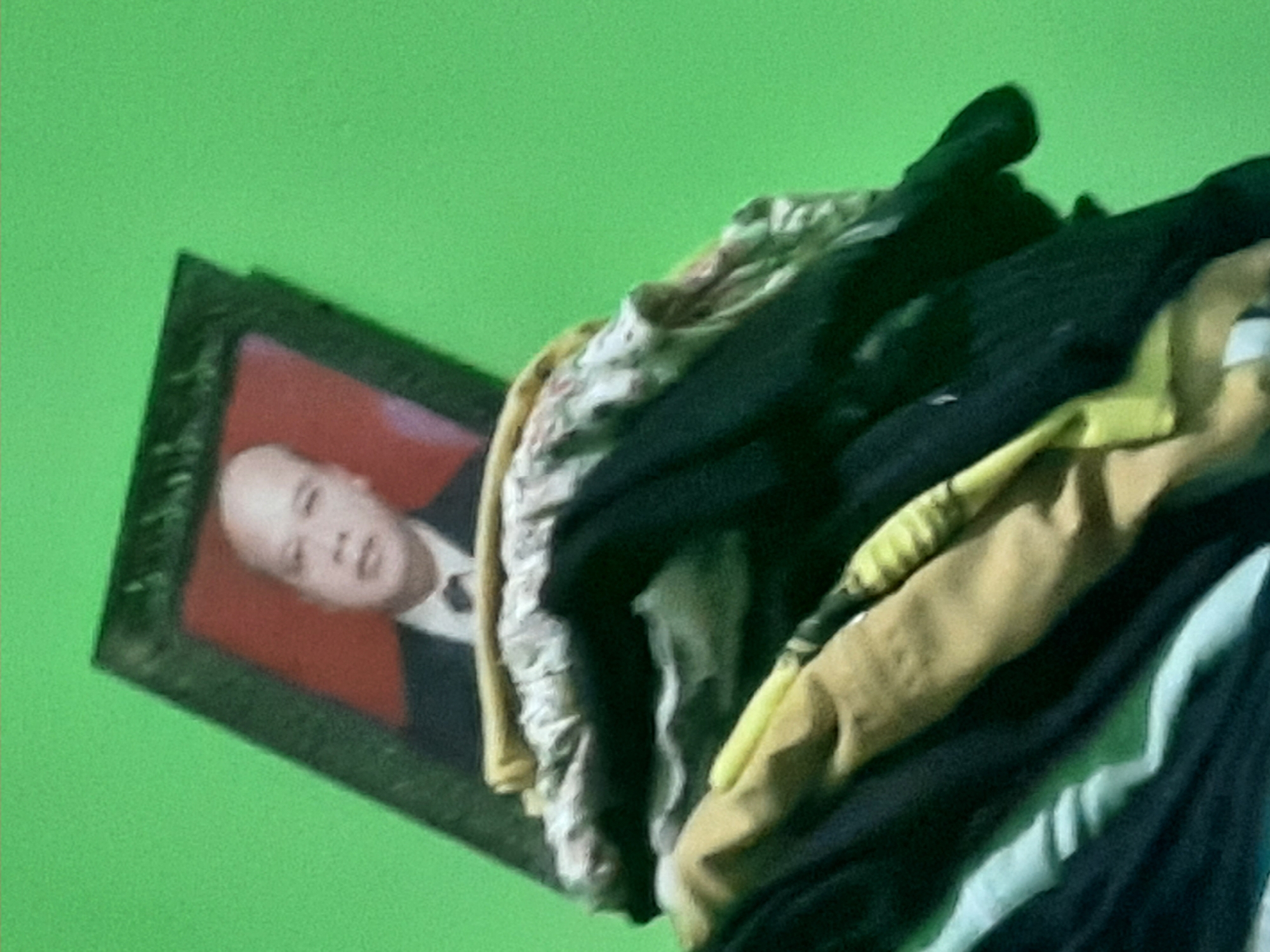 In Sumampir, Indonesia, a framed portrait of a bald man in a suit and tie is propped up against a pile of folded clothing. The portrait's background is a solid red. The clothing is stacked high and appears to be a mix of fabrics and colors, including light yellow, dark blue, and a patterned fabric. The background behind the portrait and clothing is a solid, bright green.