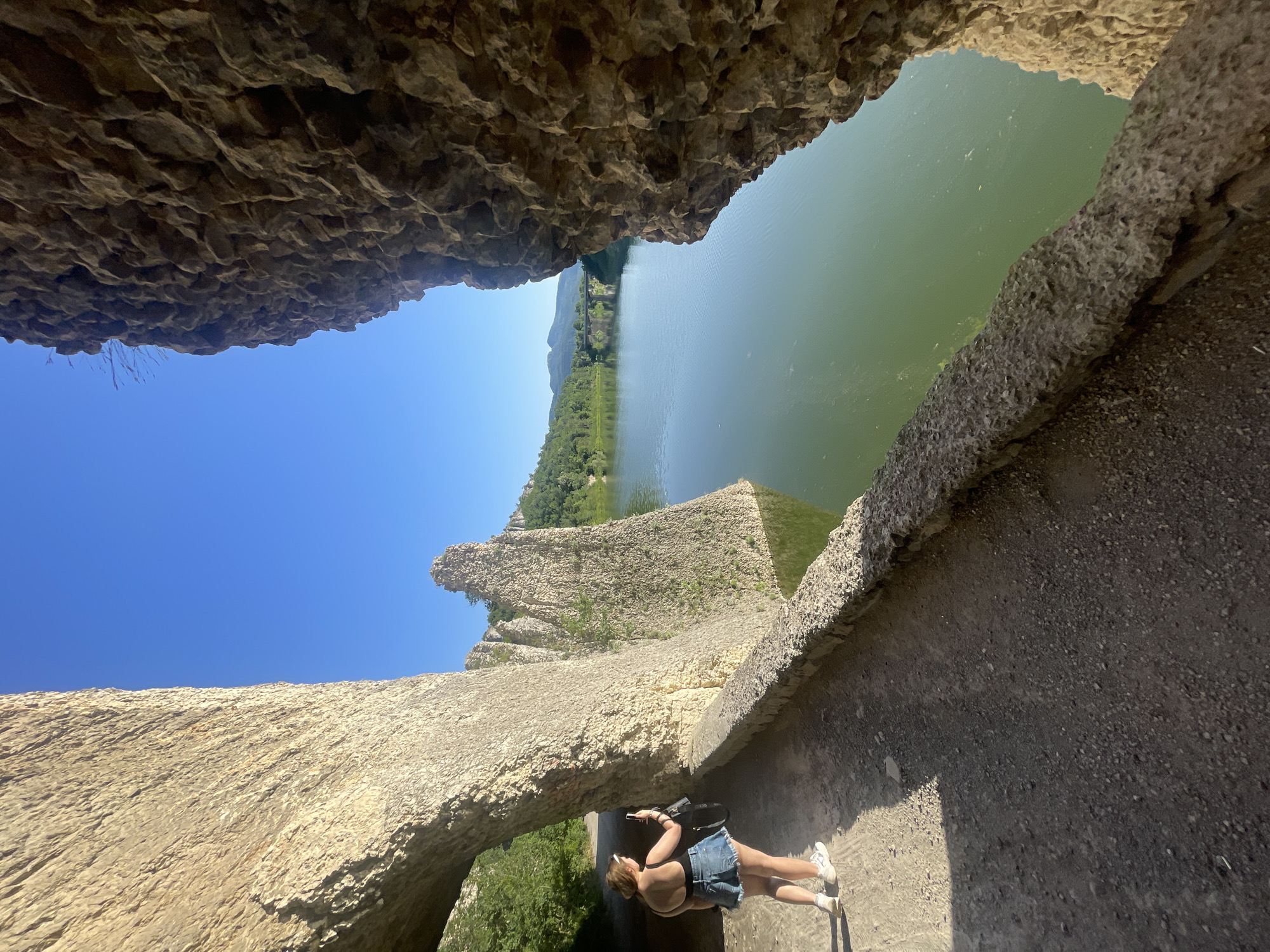 In the city of Asparuhovo, Bulgaria, a woman stands on a stone pathway, taking a photograph with her phone. She is wearing a black tank top and denim shorts, with white sneakers. The pathway is carved into a natural rock formation that arches overhead on the left. Ahead, the pathway runs alongside a body of calm green water, bordered by a low stone wall. To the right of the pathway and water, more natural rock formations rise vertically. In the distance, across the water, a bridge spans the width of the frame, with hills and mountains visible beyond. The sky is clear and blue.
