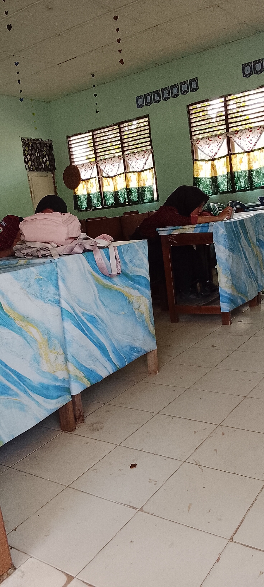 The image depicts the interior of what appears to be a classroom or meeting room in Jepara, Indonesia. Two individuals, likely students, are visible. One is lying face down on a table, head resting on their arms, while the other is leaning forward with their head down on a table, possibly asleep or concentrating. Both individuals are wearing modest clothing, with one wearing a hijab and the other a dark head covering.
The room is decorated with heart-shaped ornaments hanging from the ceiling and along the walls. A banner with the word "WELCOME" is also affixed to the wall. Two large windows with slatted blinds are present, revealing glimpses of greenery outside. The windows are adorned with curtains featuring a floral pattern in green, yellow, and white.
The tables in the foreground are covered with blue and white patterned cloths, with the pattern resembling marble or swirling waves. The floor is tiled with light-colored square tiles, showing some signs of wear and dust. The overall atmosphere suggests a casual or relaxed setting, possibly during a break or a less formal class session. The lighting appears to be natural, coming from the windows, and possibly supplemented by overhead lighting.