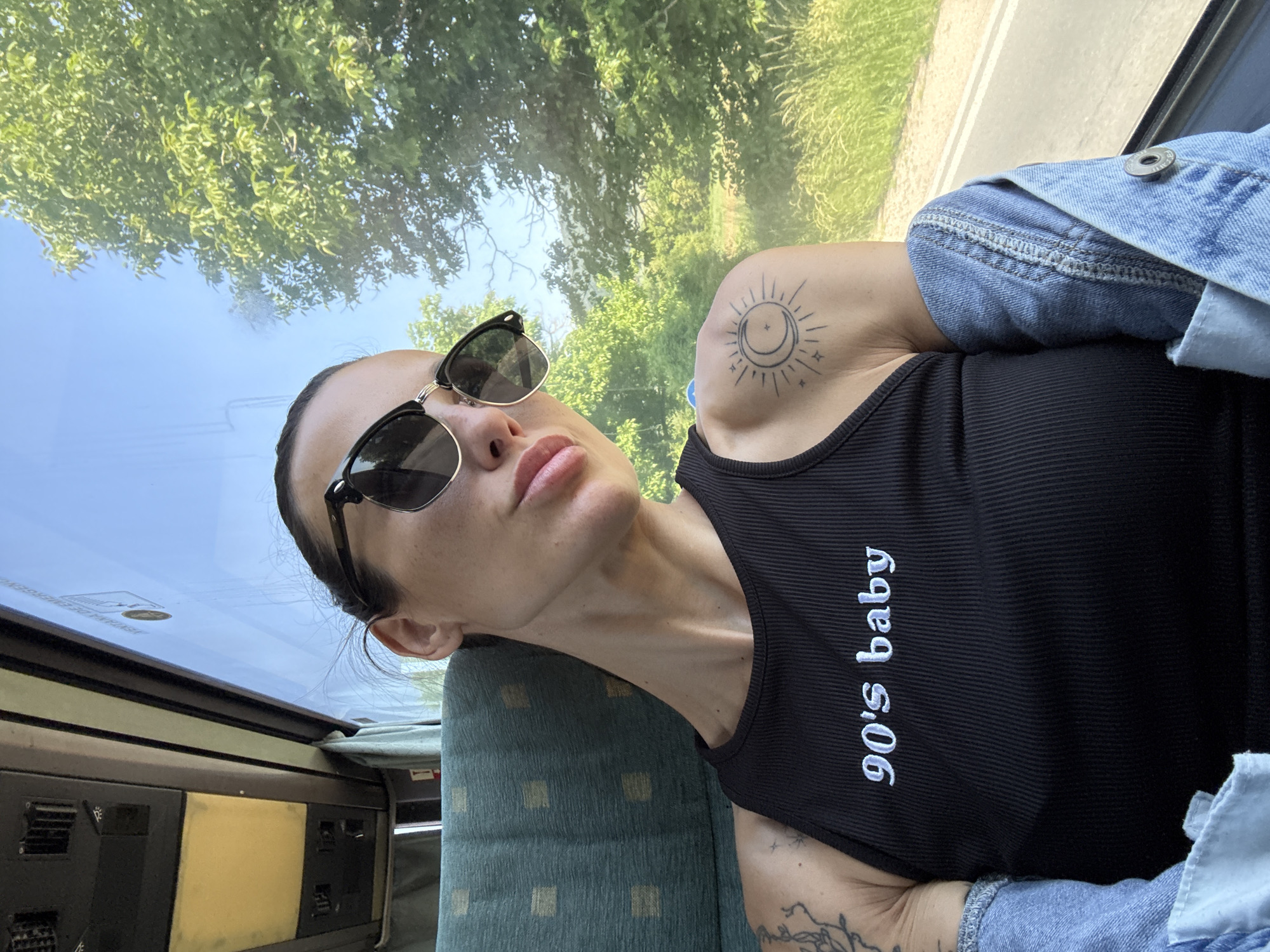 A woman wearing sunglasses and a black ribbed tank top with the words "90's baby" on it is sitting on a patterned seat in a vehicle, possibly a bus or train, in Riahovtsite, Bulgaria. She has a denim jacket draped over her shoulder and tattoos visible on her arms. The window shows trees and foliage passing by.