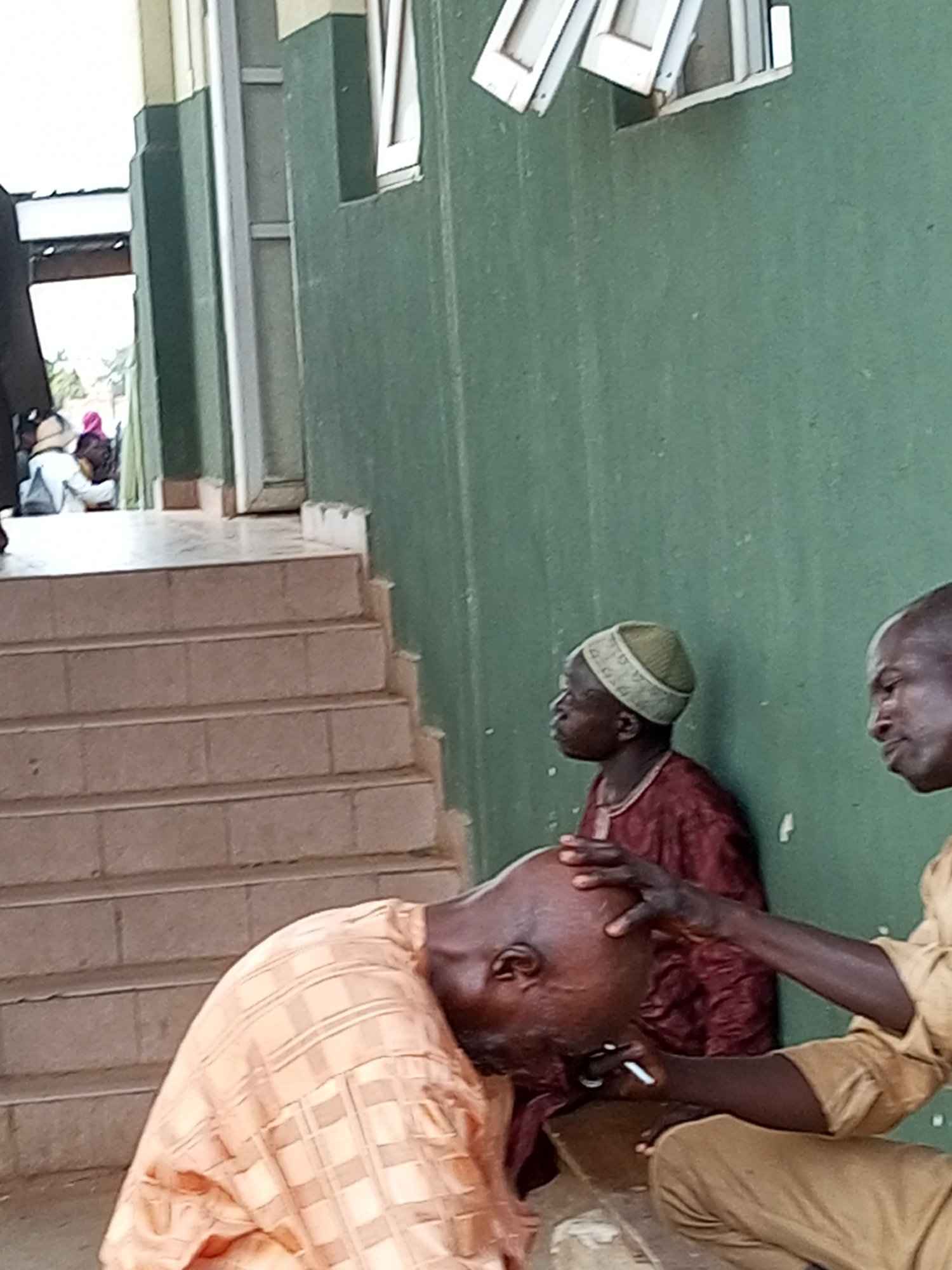 The image depicts an outdoor scene in Gombe, Nigeria, likely on a sunny day given the lighting. In the foreground, a man is having his head shaved by another man. The man receiving the haircut is wearing an orange and brown checkered traditional outfit and is bent forward. The barber, dressed in a tan outfit, is using a razor to shave the man's head. His hand is also resting on the shaved head.
In the middle ground, another man is seated, facing left and looking away from the camera. He is wearing a maroon garment and a green cap. He appears to be observing the haircutting activity or is simply present in the scene. The background shows a green wall with some architectural elements, possibly part of a building. To the left, there are steps leading up to an entrance area where several other people, including women wearing headscarves, are visible in the distance. The overall setting suggests a public space or the exterior of a building in a community area. No visible text is present in the image. The atmosphere appears calm and focused on the task at hand.