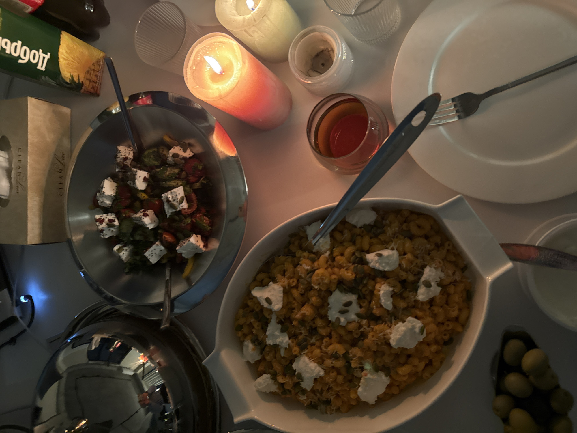 A tabletop illuminated by candlelight displays a meal. The table is set with a white plate and a fork. Next to the plate is a glass containing a reddish liquid. A bowl holds pasta topped with what appears to be cheese. A second, smaller bowl has a salad with what could be feta cheese, tomatoes, and greens. A covered serving dish sits adjacent to the salad bowl. Nearby are olives, and a small bowl with a white sauce. A carton of pineapple juice is also visible. The setting is suggestive of a dining experience. The location in Yerevan, Armenia, is not ascertainable from the image.