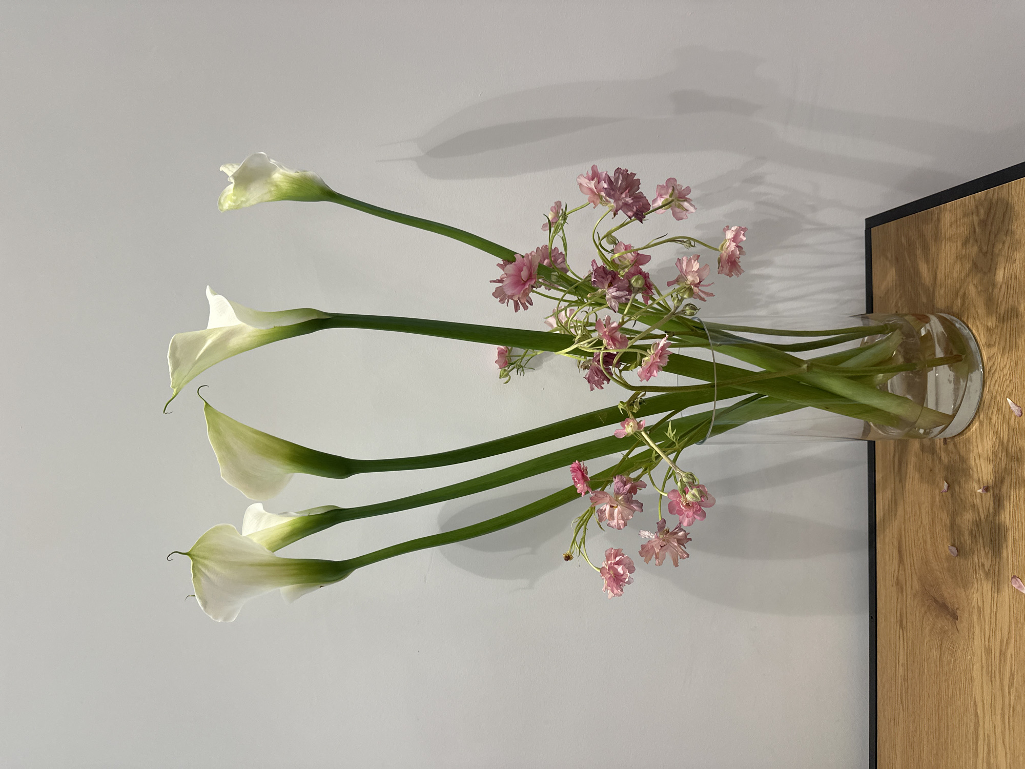 The image depicts a vase of white calla lilies and small pink flowers. The vase is cylindrical and transparent, filled with water. It sits on a wooden surface with a dark trim against a plain white wall. Some fallen petals are scattered on the wooden surface. The scene has a simple, elegant aesthetic. Location in Milan, Italy is not indicated in the image.