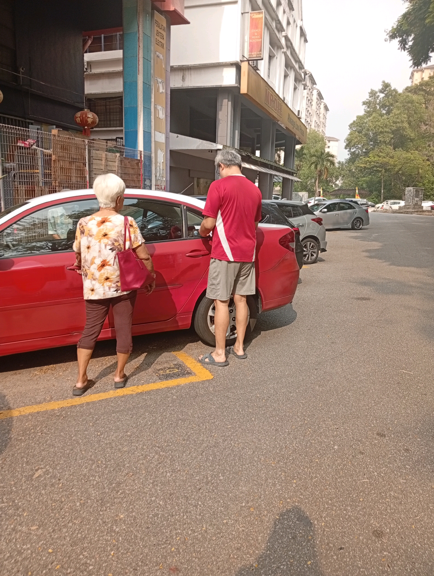 In a street parking area in Selayang Municipal Council, Malaysia, a man and an elderly woman stand beside a red car. The man, wearing a red t-shirt and grey shorts, faces the car with his back to the camera. The woman, with short white hair, is in profile, looking towards the car and holding onto its door handle. She is wearing a floral print shirt and brown cropped pants. A red handbag is slung over her shoulder. The background features a multi-story building with various shops and businesses, including one with a sign for "MIRADAY" and "Always Low Prices." Trees and other vehicles are visible in the distance. The ground is paved asphalt, marked with yellow parking lines.
