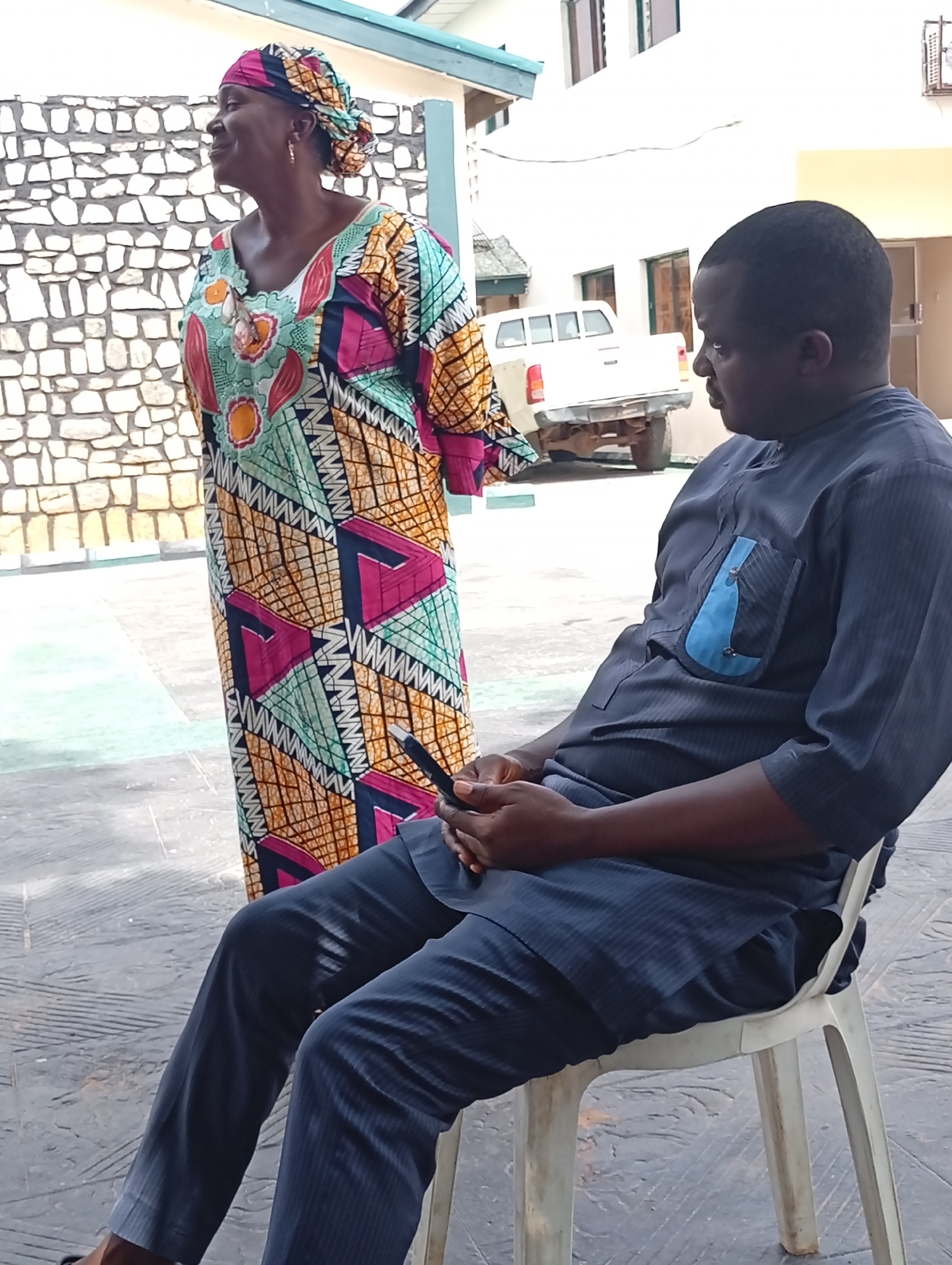 The scene appears to be outdoors, potentially within the city of Dadi Riba, Nigeria. An African woman wearing a colorful, patterned dress and head wrap stands near a stone wall. To the right, a man is seated on a white plastic chair, looking towards the woman. He is wearing a dark blue outfit with a lighter blue pocket detail. He holds an object in his hands. A white pickup truck is parked in the background. The ground is paved with a patterned surface. The buildings are painted white.