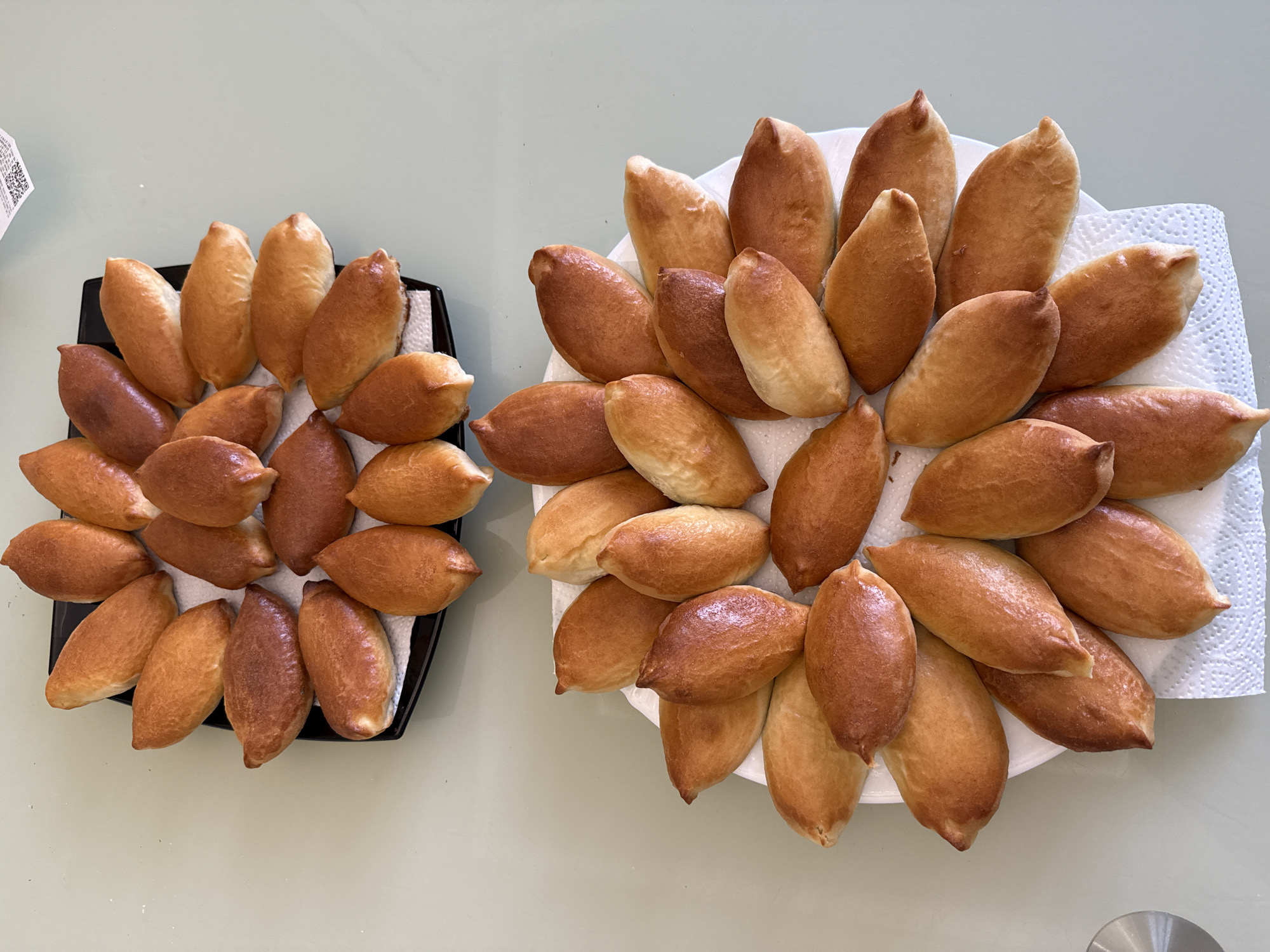 Two plates of freshly baked, oblong pastries (likely pirozhki) are displayed on a light-colored surface, possibly a kitchen countertop or table. The larger, round white plate on the right, lined with a white paper towel, holds numerous golden to light brown pastries arranged in a circular, radiating pattern. The smaller, rectangular black plate on the left, also lined with a white paper towel, contains fewer pastries, which exhibit a greater range of browning, from golden to darker brown, and are arranged more compactly.
All pastries are uniformly torpedo-shaped with slightly pointed ends. The scene is brightly lit, suggesting an indoor setting during the daytime. A small, partially visible QR code or barcode label is seen in the top-left background. A metallic object is partially visible in the bottom-right corner. The image captures food prepared for consumption in Votkinsk, Russia.