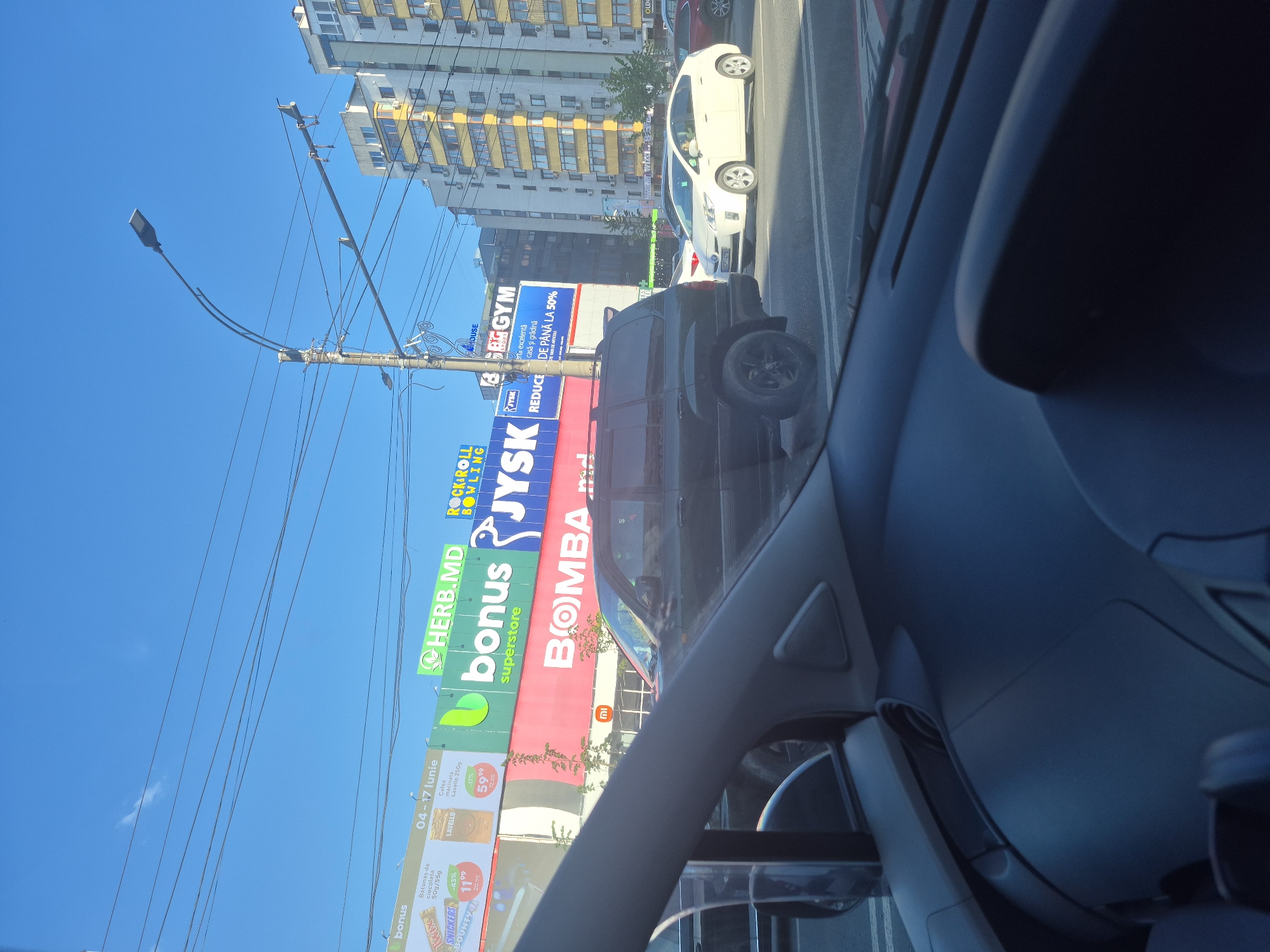 The image was taken from inside a car looking out at a street scene in Chișinău, Moldova. A large billboard displays advertisements for "HERB.MD", "bonus superstore", "JYSK", and "BOMBA". The "BOMBA" advertisement includes the logo for "MI". Another billboard promotes "ROCK & ROLL BOWLING". A street lamp and utility poles with wires are visible against a blue sky. Buildings of varying heights and architectural styles line the street, with one building prominently featuring yellow and white facades. Traffic is visible, including a dark SUV and a white car. The overall scene depicts an urban environment with commercial advertising.