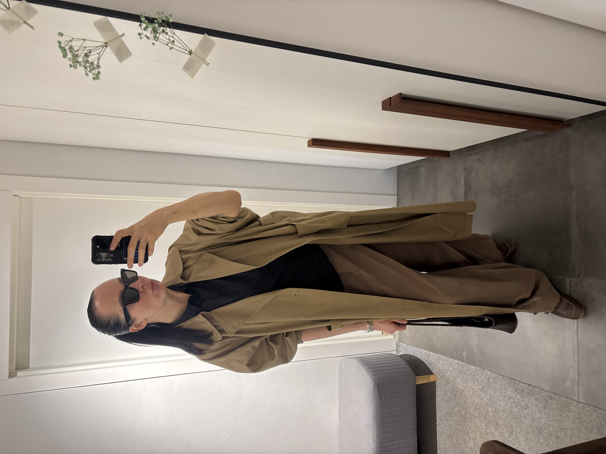 A woman is captured in a mirror selfie indoors, standing and holding a black smartphone with her right hand to photograph her reflection. She is positioned at a slight angle, wearing large, dark rectangular sunglasses that obscure her eyes. Her dark hair is pulled back, revealing her profile. She is dressed in a light brown or khaki trench coat layered over a black top and loose-fitting, wide-leg brown pants. Her footwear consists of brown shoes, and she holds a black woven handbag in her left hand, which also displays a silver-toned watch on her wrist.
The background reflects a modern and minimalist interior. The white ceiling features delicate decorative elements: several small bunches of green leaves, possibly baby's breath, are taped with white adhesive strips to a thin string or wire running horizontally. Further along the ceiling, two rectangular wooden beams are mounted. The walls appear to be light, transitioning to a darker gray, possibly concrete, on the lower right side. A gray upholstered bench with a wooden leg is partially visible on the left, and the floor is a uniform gray. The lighting is bright and even, indicative of an indoor setting. No discernible text is visible in the image. The scene suggests a retail environment or a contemporary personal space in Chaikovsky, Russia.