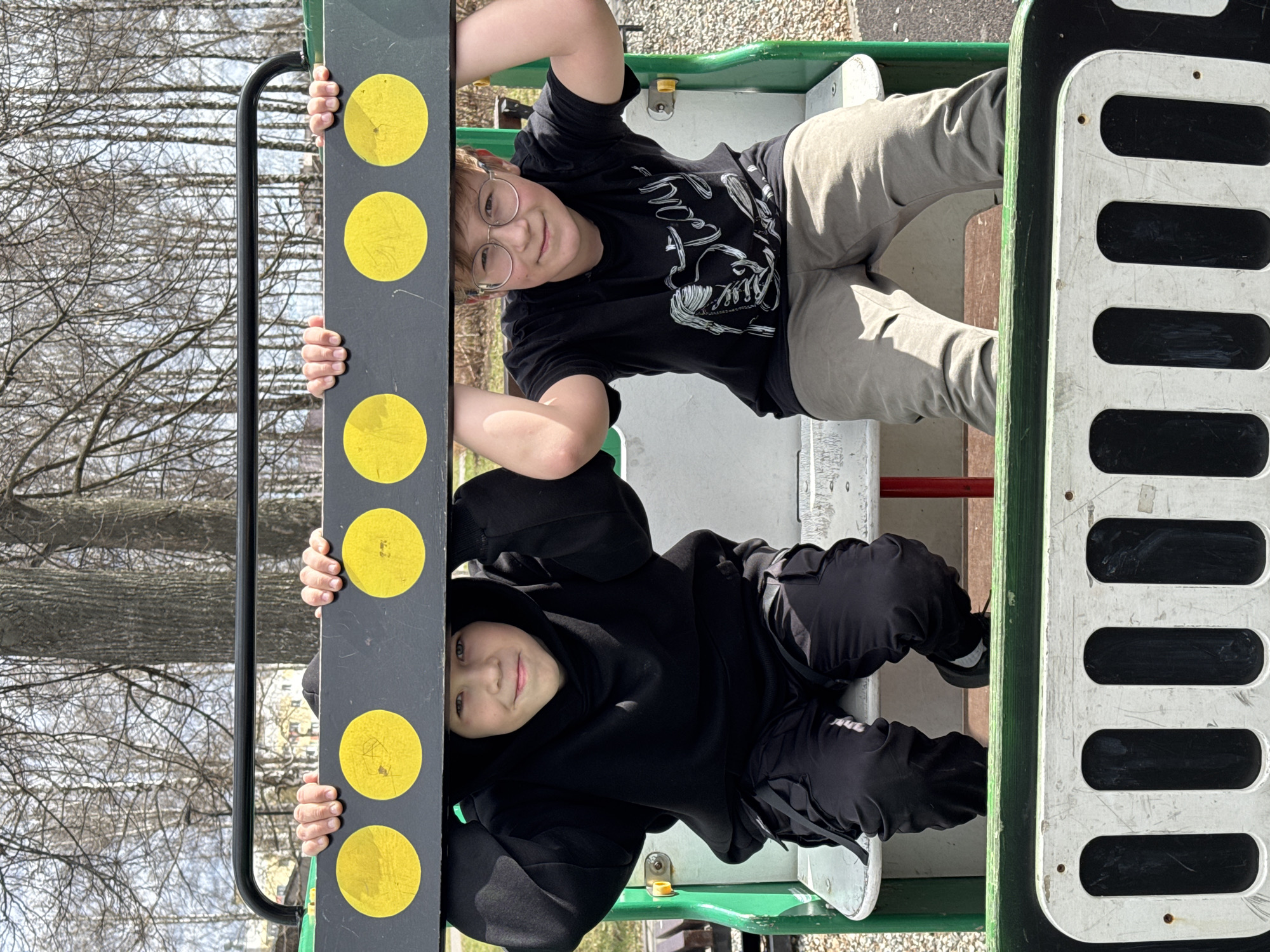 Two children are on playground equipment. The equipment appears to be part of a train. One child, wearing glasses and a black t-shirt, is above the other. The child below wears a black hooded sweatshirt. Both are holding onto a black vertical piece with yellow circles. The background shows trees without leaves and a light-colored ground surface. The location is Zav'yalovo, Russia.