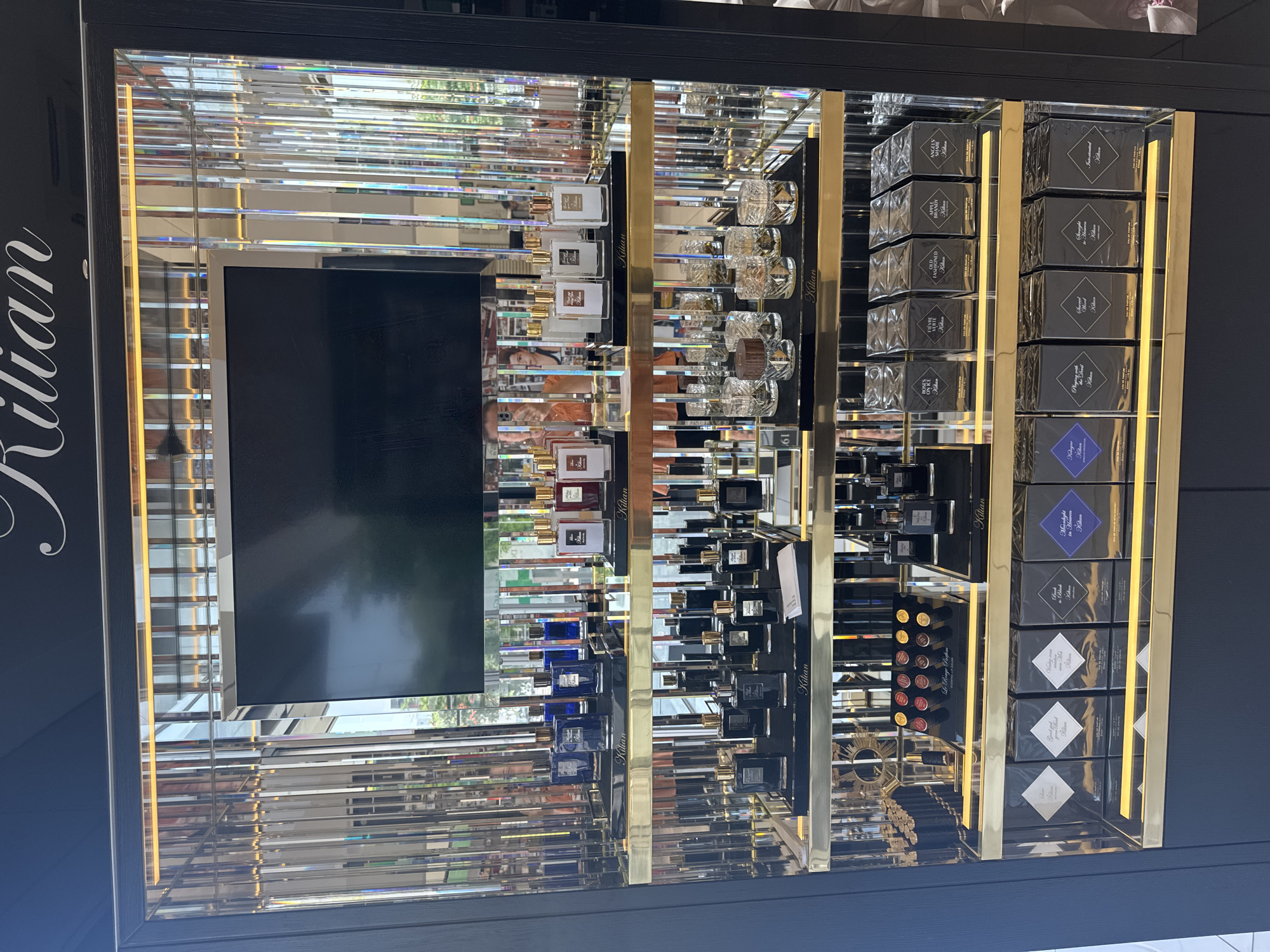 The image shows a retail display of Kilian perfumes in Germasogeia Municipality, Cyprus. The display features multiple shelves filled with various perfume bottles and boxed sets. The shelves are illuminated by warm lighting, and the background consists of mirrored panels that reflect the products and the surrounding environment. A black screen is mounted at the top of the display. On the shelves, there are rows of distinctively shaped perfume bottles in shades of blue, white, and dark glass, some with gold caps. There are also decorative glass vessels and a selection of lipstick shades arranged on a black tray. At the bottom, several boxed perfume sets are neatly arranged. The overall impression is of a high-end retail presentation.