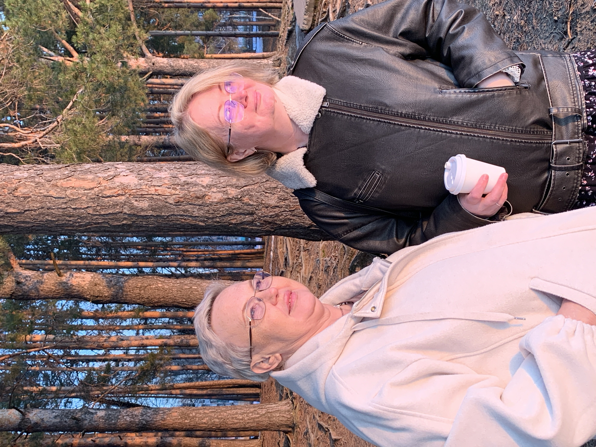 Two women are posing outdoors in front of a backdrop of trees and what appears to be a wooden structure or fence. The location is in Chaikovsky, Russia.
The woman on the upper right is wearing a black leather jacket with a light-colored faux fur collar and a belt. She is holding a white cylindrical object that may be a travel mug.
The woman on the lower left is wearing a light-colored hooded jacket or coat. She has short, greyish hair and is also wearing glasses.
The trees in the background appear to be pine or evergreen trees. The wooden structure behind them looks like logs arranged horizontally. The overall lighting suggests it could be late afternoon or early evening.