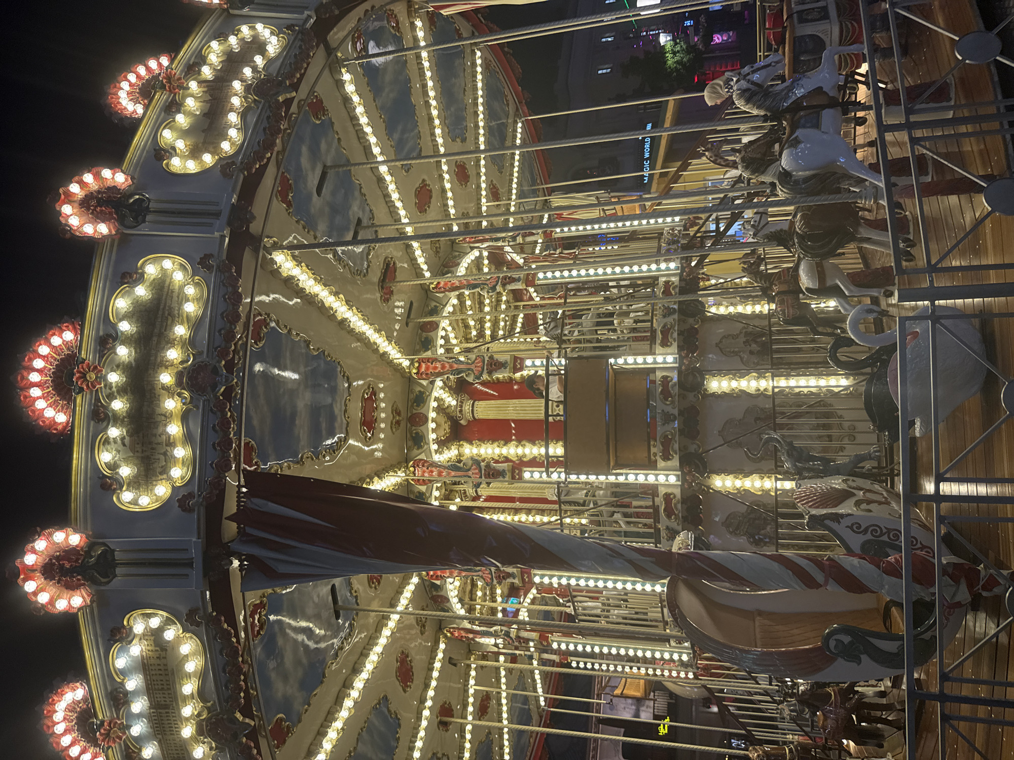 The image shows a carousel at night, illuminated by numerous lights. The carousel is multi-tiered and decorated with ornate designs and painted scenes. Several horses and swan-shaped seats are visible on the lower level. In the background, a building with "MAGIC WORLD" signage is partially visible, suggesting this location is an amusement or entertainment district. The overall scene depicts a festive and lively atmosphere common to carnivals or fairgrounds.