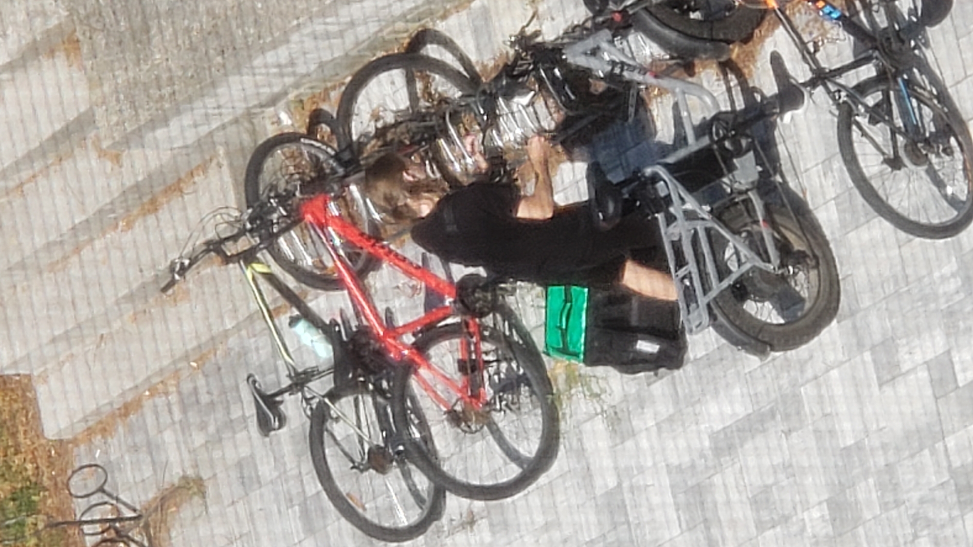 A group of bicycles, including a red mountain bike and two other bikes, are parked on a tiled surface. A person in a black shirt and shorts stands next to the red bike, appearing to be adjusting something on another bicycle in the foreground. A green delivery bag is visible near the person. The background shows a set of concrete stairs. The scene appears to be an outdoor area, possibly a public space or residential complex.