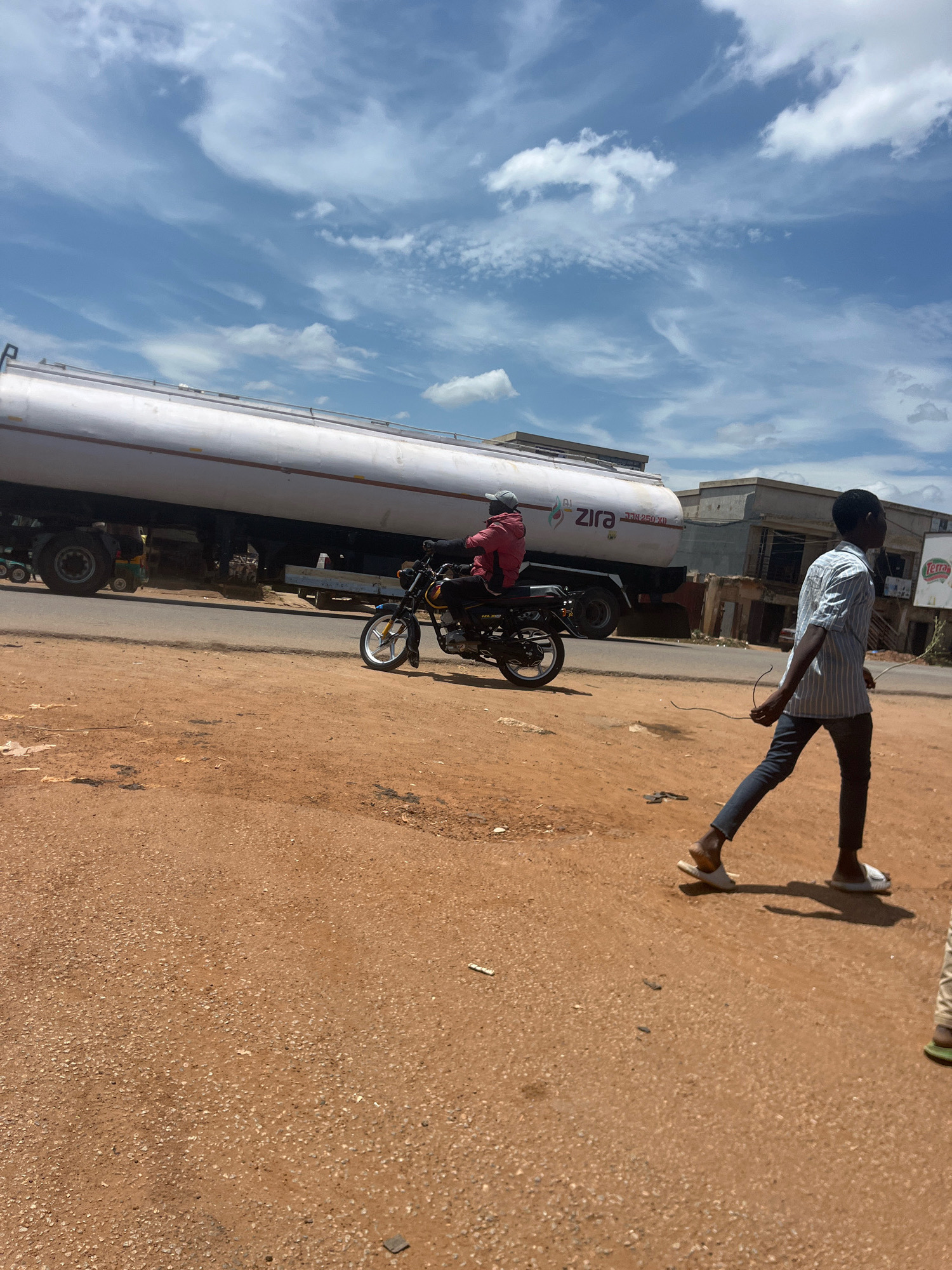 A wide shot shows a street scene in Rafin Par, Nigeria. A white tanker truck with "ZIRA" written on its side is parked on the road, as is a motorcycle with a rider wearing a red jacket and a grey helmet. A young man wearing a blue and white striped shirt and dark pants walks away from the camera on the dirt road in the foreground, carrying a thin wire. Buildings with light-colored facades are visible in the background. The sky is blue with scattered white clouds.