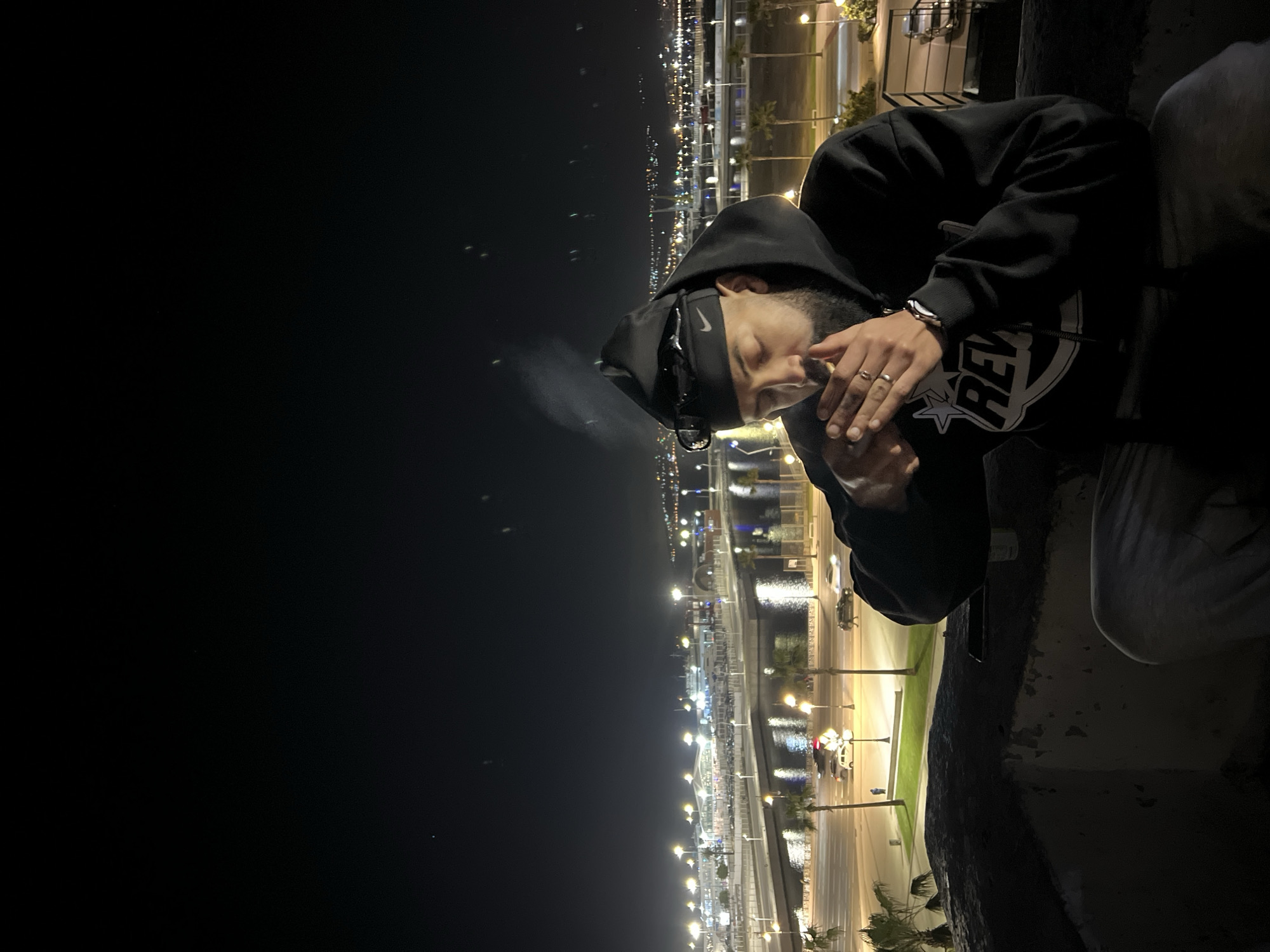 The image depicts a nighttime urban scene from an elevated vantage point in Tanger, Morocco, specifically overlooking the Tanger Ville port area.
In the foreground, a bearded male figure is seated on a weathered concrete ledge, visible from the chest up. He wears a black hoodie with a white "REV" graphic, a black beanie featuring a white Nike swoosh, and black sunglasses pushed onto the beanie. He holds a lit cigarette to his mouth with both hands, from which a plume of smoke is visible ascending. Rings are present on his left hand, and a watch on his left wrist. A mobile phone and a white cup are positioned on the ledge near him.
The background displays the illuminated infrastructure of the Tanger Ville port. A multi-lane road, bordered by streetlights and palm trees, runs horizontally across the midground, with several moving vehicles discernible. Beyond the road, a body of water, likely the harbor, reflects the artificial lights. Across the water, brightly lit port facilities, including what appears to be a ferry terminal with numerous structures and parked vehicles, are visible. Further in the distance, a sprawling pattern of city lights outlines an urban area across the bay under a dark, clear night sky.