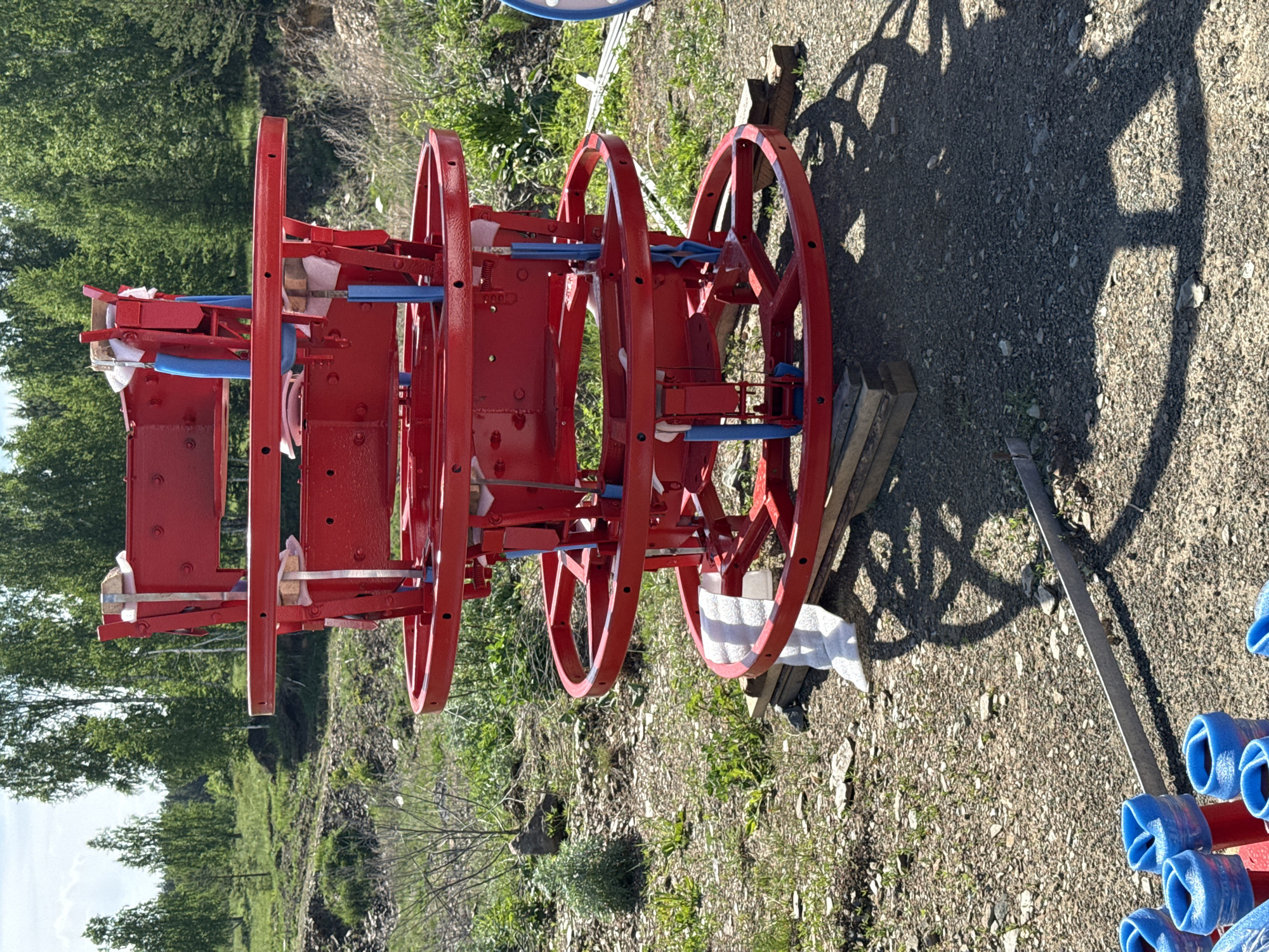 The image depicts a red metal structure with multiple wheels, possibly part of a machine, resting on a gravelly surface with vegetation in the background. Blue padding material is attached to some of the metal parts. The scene is outdoors, with trees and shrubs visible in the background, and the presence of shadows indicates a sunny day. The structure appears to be undergoing assembly or maintenance, with white cloth or padding material also present. The location is not verifiable as Leninsk, Russia, from the visual information provided.