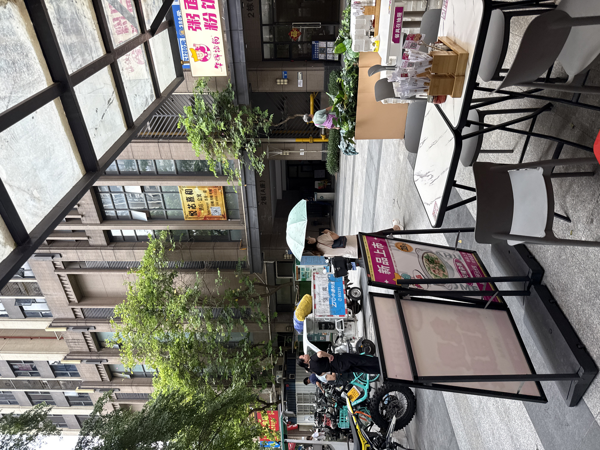The image depicts an outdoor scene in front of a modern building with multiple floors and windows. In the foreground, there are two tables with chairs, set up for dining. One table has a marble-like surface and is adorned with chopsticks in wooden holders and a small jar. The other table is partially visible. Several A-frame signs are positioned on the pavement; one sign features an image of a bowl of food and text in Chinese. To the left, a variety of parked motorcycles and scooters are visible. A white delivery truck with blue and yellow branding, including the text "ZTO," is present behind the parked vehicles. In the background, several people are present. One woman, holding a light green umbrella, stands near the delivery truck. Another woman is seen walking away from the truck. The building in the background has a beige facade with vertical and horizontal architectural details and numerous windows. Green trees and foliage are interspersed throughout the scene, suggesting an urban environment with some greenery. The overall impression is a commercial or residential street in a city.