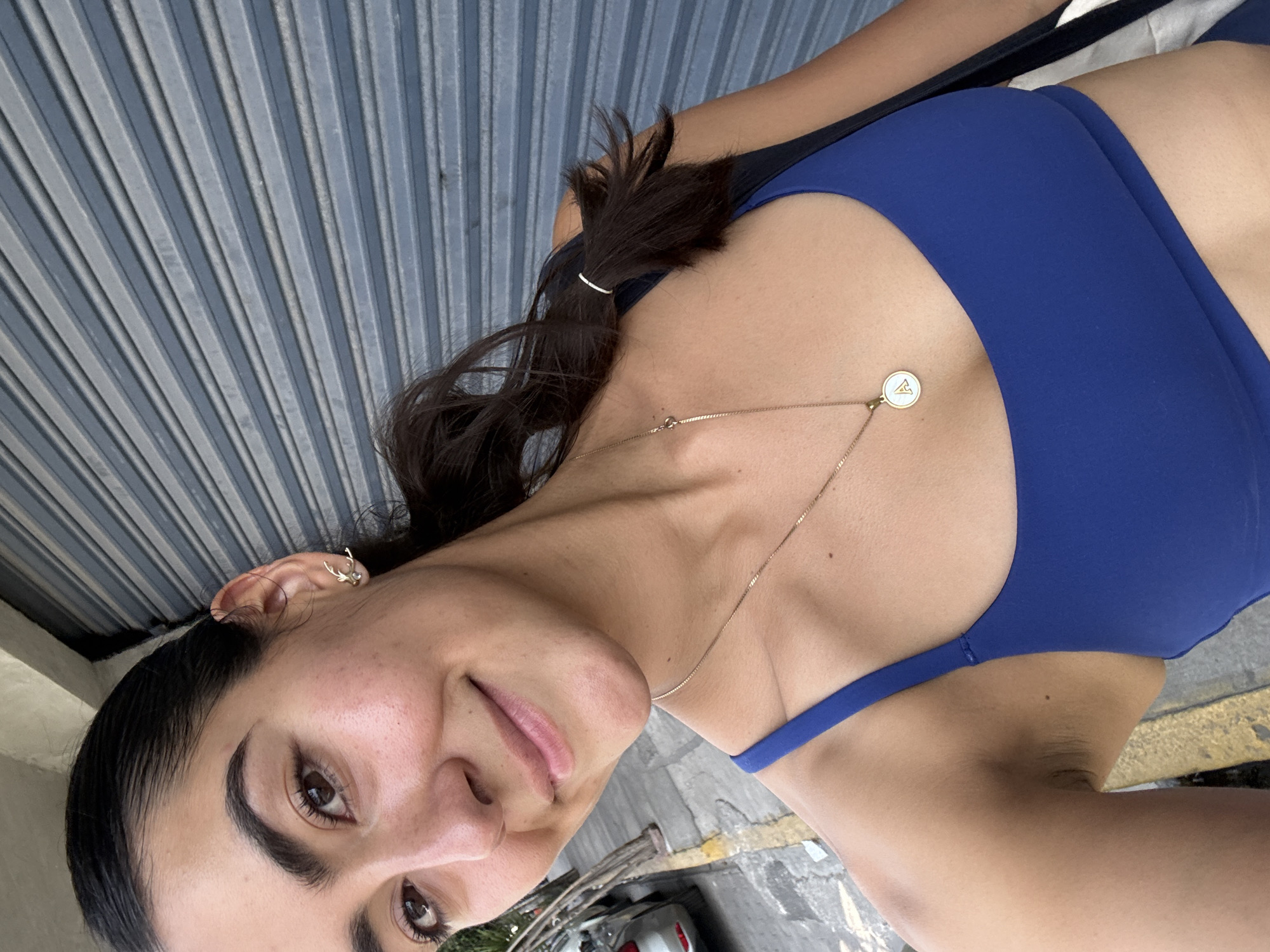 A woman with dark hair, wearing a blue sports bra and a gold necklace, is posing for a picture. She is standing outside in an urban setting, with a textured grey wall behind her. She appears to be in San Andrés Cholula, Mexico.