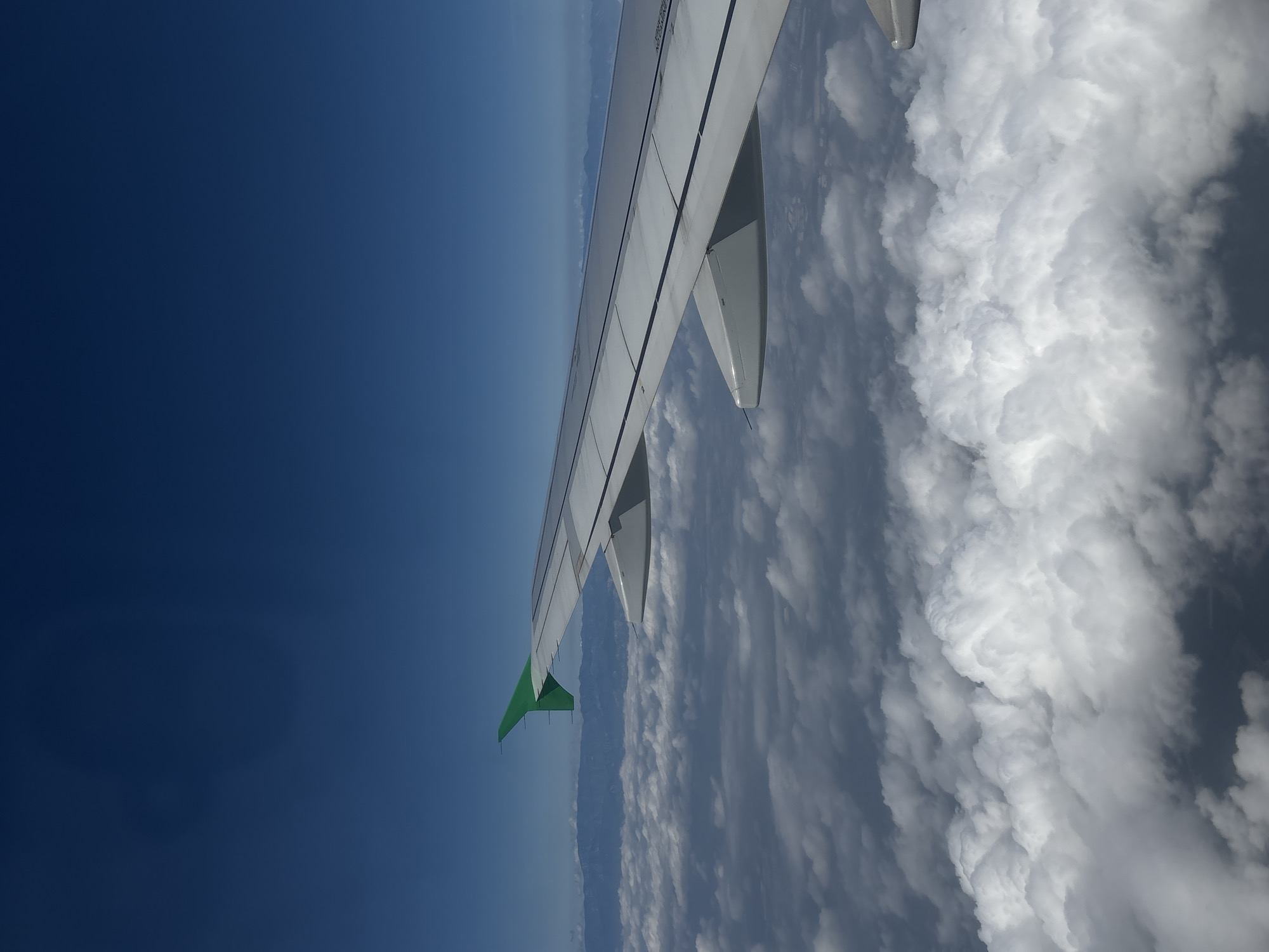 The image shows an aerial view of an airplane wing with a green tip, extending over a landscape of clouds and mountains. The sky is a deep blue, indicating a clear day. The wing is white with dark gray panel lines, and a portion of the airplane's fuselage is visible. Below the wing, fluffy white clouds stretch across the horizon, obscuring the ground below. In the distance, a range of mountains can be seen peaking through the clouds. The scene appears to be during daylight hours, likely in the afternoon given the lighting. The perspective suggests the photo was taken from a passenger's window seat on a flight. There is no visible text in the image.