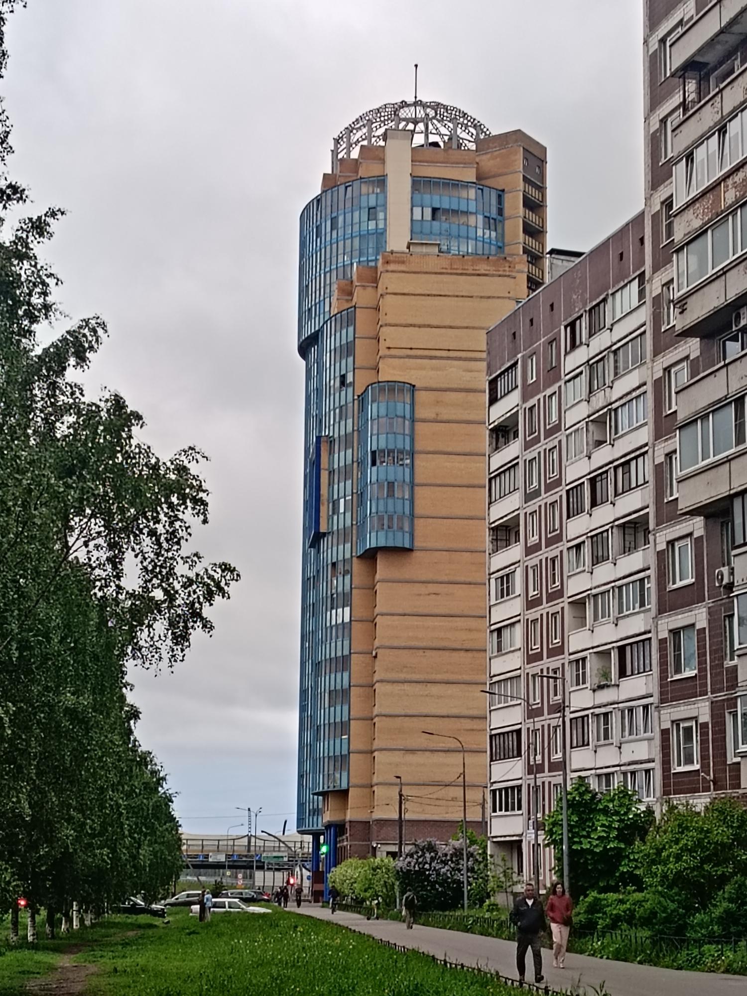 The image depicts a street scene in Saint Petersburg, Russia, with a mix of modern and older architectural styles. On the right is a tall apartment building with a repetitive pattern of windows and balconies. To the left of the apartment building is a more contemporary, multi-tiered structure with rounded glass sections and brickwork. It features a decorative open structure on top.
A tree line borders the left side of the image, with green grass and a walking path visible in the foreground. Pedestrians are walking along the path. Additional cars and infrastructure such as streetlights and overpass can be seen in the background. The sky overhead is overcast.