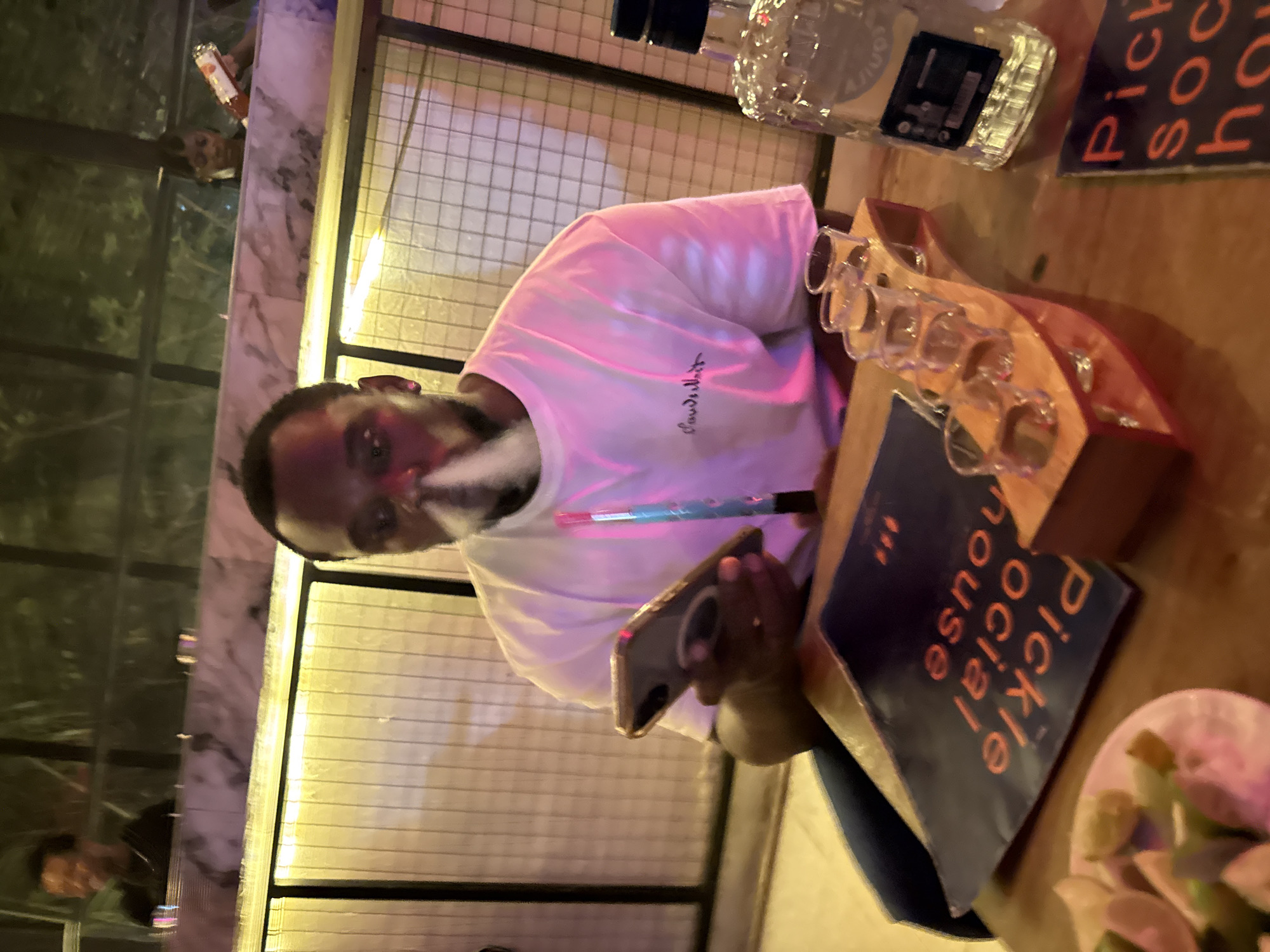 This media file shows a man in a bright pink t-shirt, holding a phone and a vape pen. He appears to be smoking from the vape pen, with a cloud of vapor visible. Behind him, several shot glasses are arranged on a wooden rack. A bottle of what looks like liquor is also visible on a wooden table. A book with the text "pickle social house" in orange letters on a dark blue cover is placed next to the shot glasses. The man has a thoughtful or slightly serious expression on his face. The lighting suggests it could be nighttime or a dimly lit indoor setting, possibly a bar or restaurant. The background shows a mesh-like metal fence and some reflective surfaces, hinting at a modern or industrial-style venue. There's also a small plate with what appears to be food items, possibly garnishes like lime wedges, in the lower right corner of the image. The overall scene suggests a relaxed social gathering.