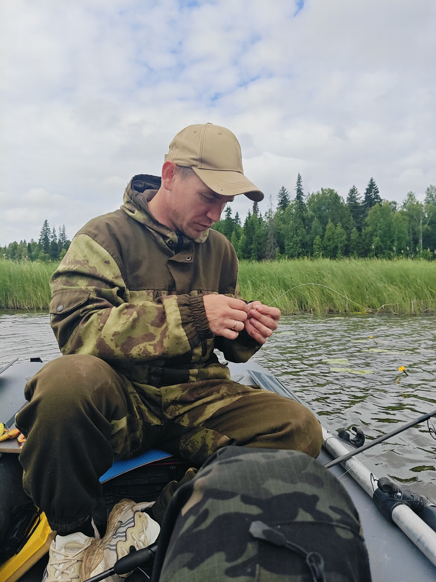 A man wearing a camouflage jacket and a tan baseball cap is sitting in an inflatable boat on a body of water. He is focused on his hands, which are holding fishing line and appear to be tying a knot. The boat is on a lake or river, with tall reeds and trees visible in the background. The sky is overcast with clouds. Fishing rods are present in the boat, and yellow fishing floats are visible in the water. The man is wearing sneakers. The image captures a moment of preparation for fishing.
