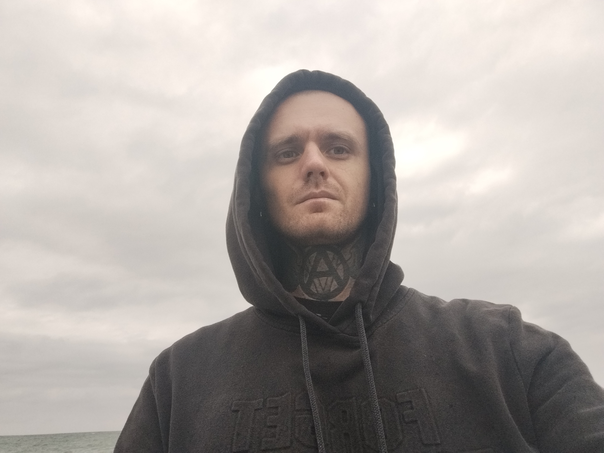 A man in a dark hooded sweatshirt is looking directly at the camera. He has a tattoo of an anarchy symbol around his neck. The background is a cloudy sky and a body of water. The scene appears to be a coastal area. The location is specified as the city of Batumi, Georgia.