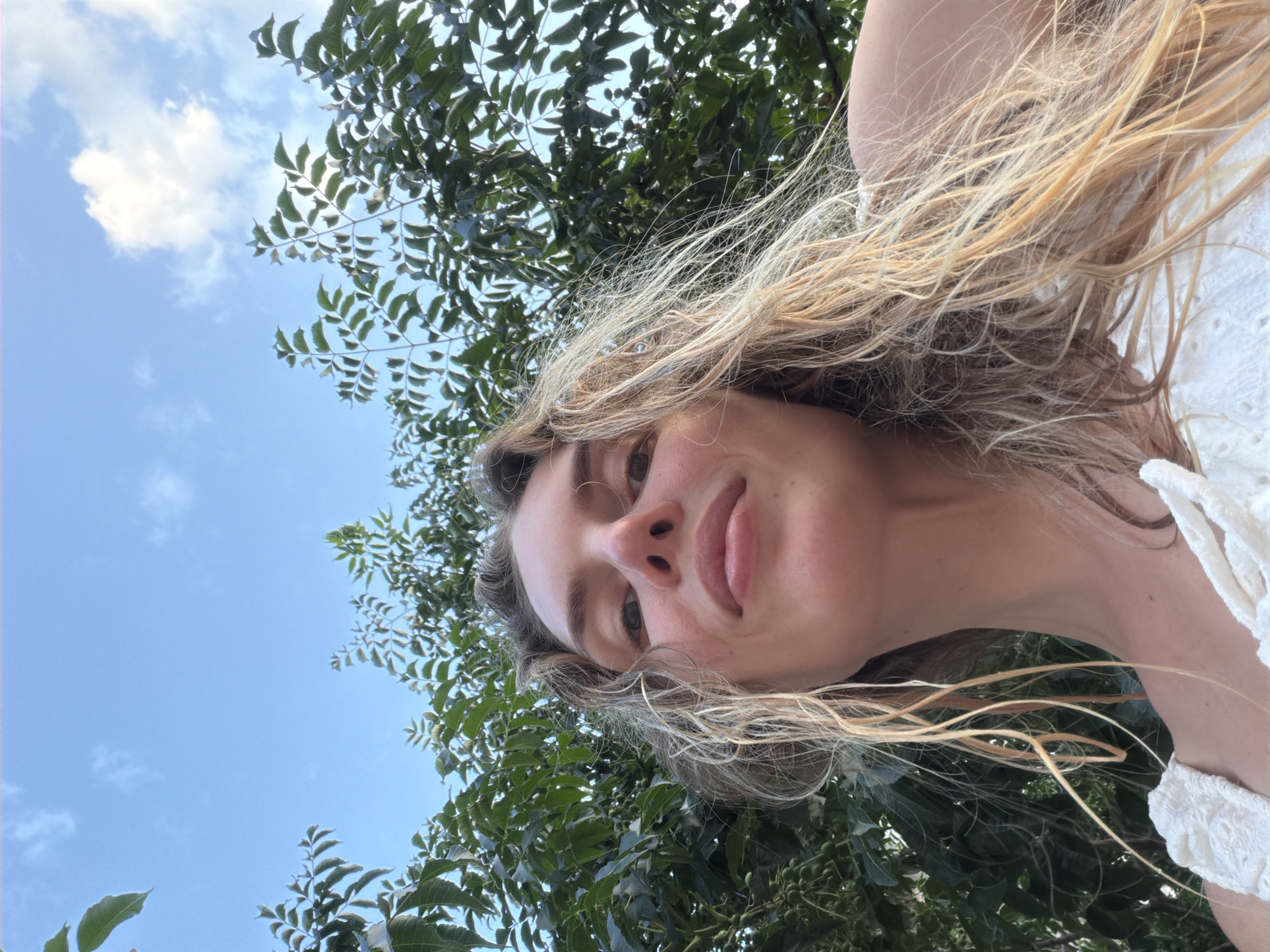A low-angle, close-up, point-of-view shot of a woman's face is captured outdoors against a backdrop of lush green foliage and a bright blue sky with scattered white clouds. The woman has long, wavy blonde hair that is slightly damp and falls around her face and shoulders. She is wearing a white top with intricate detailing around the neckline. Her eyes are open, and she is looking slightly upwards, with a gentle smile on her face. The focus is on her features, with the background foliage blurred. The scene appears to be set in a natural environment, possibly a garden or park, and there is no indication of the city of Budva, Montenegro in the image.