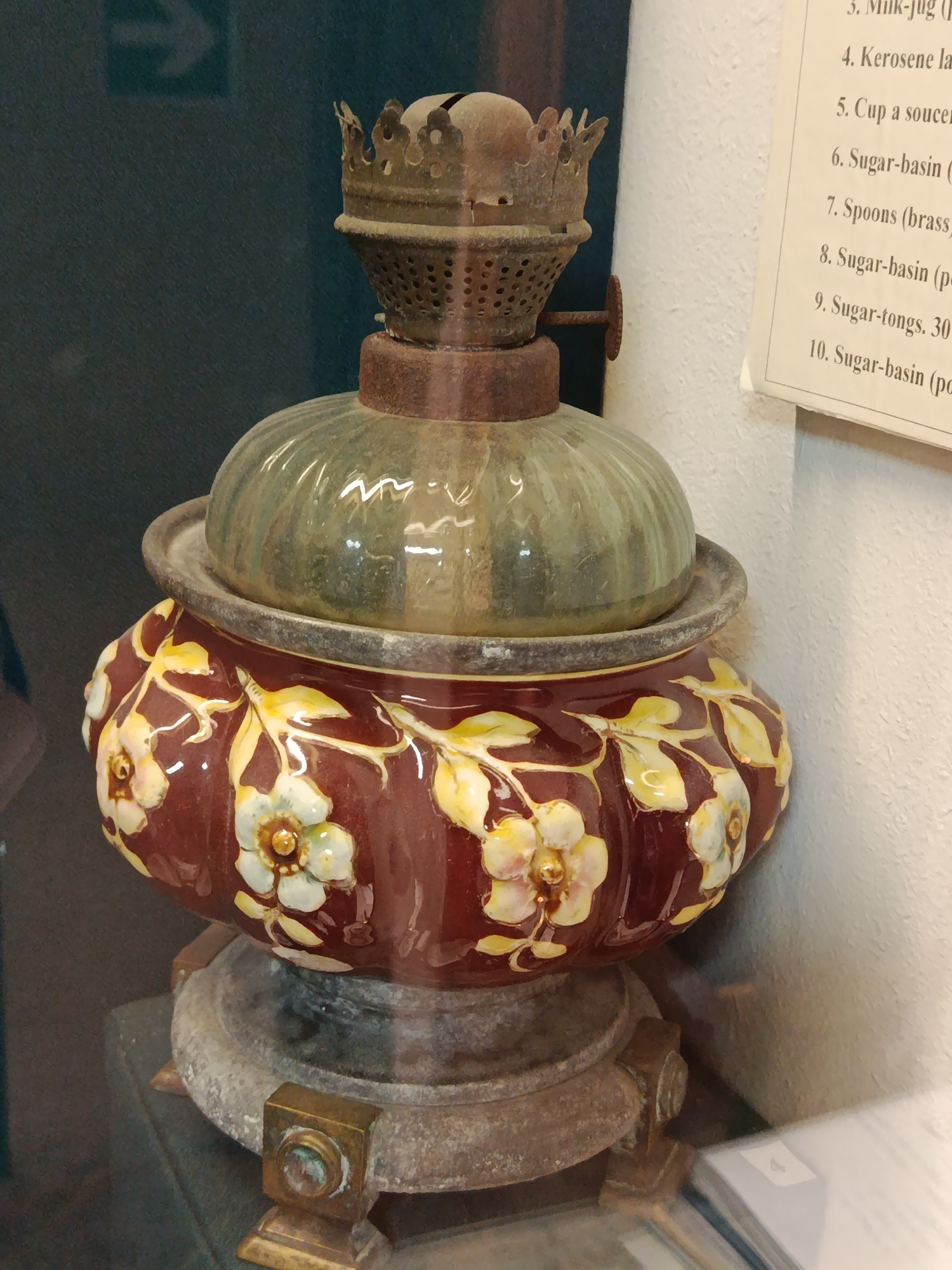 The image shows a close-up of an antique oil lamp. The lamp has a bulbous, reddish-brown ceramic base decorated with floral motifs in yellow and white. A clear, ribbed glass dome sits atop the base, and above that is a metal wick holder with a decorative, crown-like top. The lamp appears to be sitting on a weathered stone pedestal. To the right of the lamp, a portion of a white paper list is visible, with handwritten numbers and descriptions, suggesting it is part of a museum exhibit. The background on the left is dark and out of focus, showing a faint, green directional arrow on a dark surface. The lighting is diffused. The scene is likely located in a museum or historical exhibition space in Braslaw, Belarus.